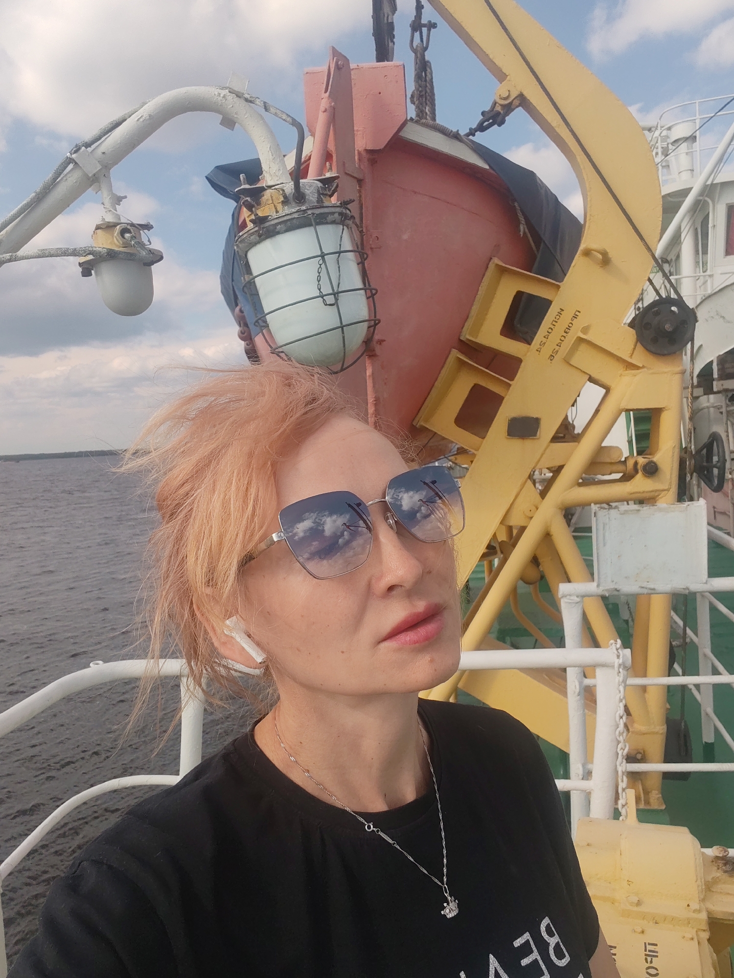 A woman with light-colored hair and sunglasses takes a selfie on the deck of a ship. She is wearing a black t-shirt and a silver necklace with a crown pendant. In the background, there is a red life raft and a yellow crane. The sky is cloudy. The image appears to have been taken in Krokhino, Russia.