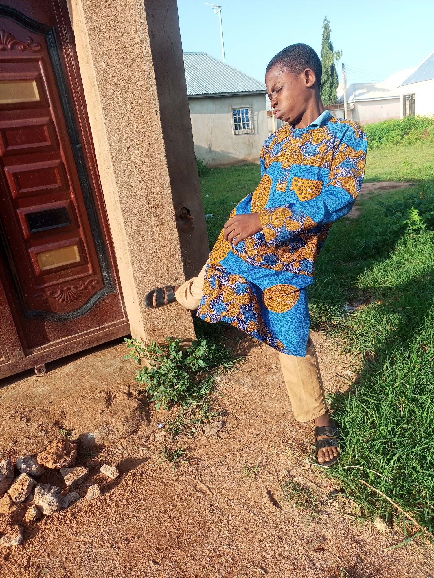A young boy stands outside a building in Tempure, Nigeria. He is wearing a blue and yellow patterned shirt and tan pants, with black sandals on his feet. The boy is looking down with his eyes squinted and mouth pursed. He is leaning against a textured brown wall next to a dark red door with decorative panels. The ground is a mix of sand and dirt, with some scattered rocks and green plants growing. In the background, there are buildings with corrugated metal roofs and green grass. The sky is clear and blue.