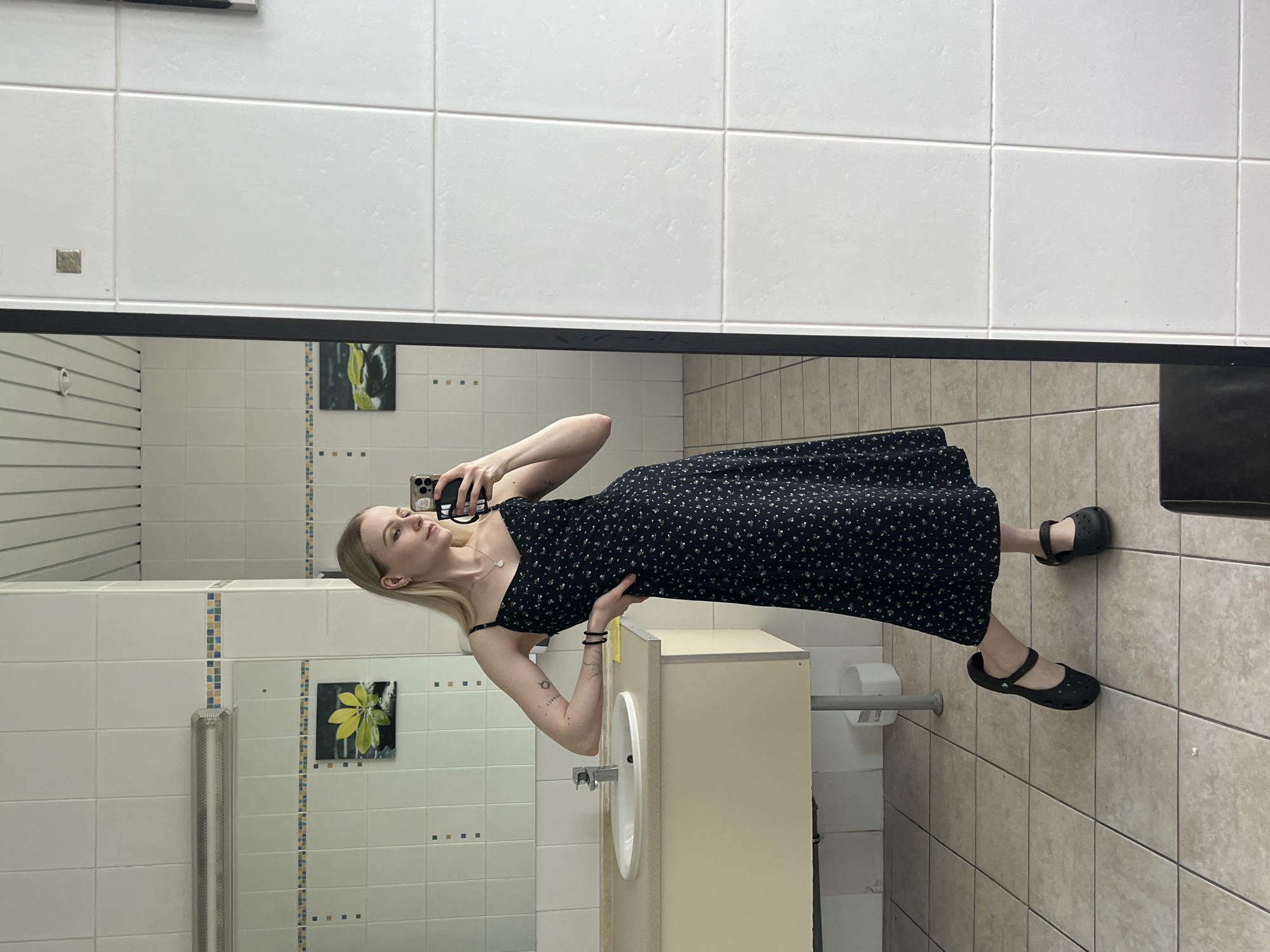 A woman is taking a selfie in a bathroom. She is wearing a black floral dress and black Croc shoes. She is standing in front of a sink and mirror. The bathroom is tiled in white and beige, and there is a picture of a yellow flower on the wall. There is no information indicating the city of Khimki, Russia.