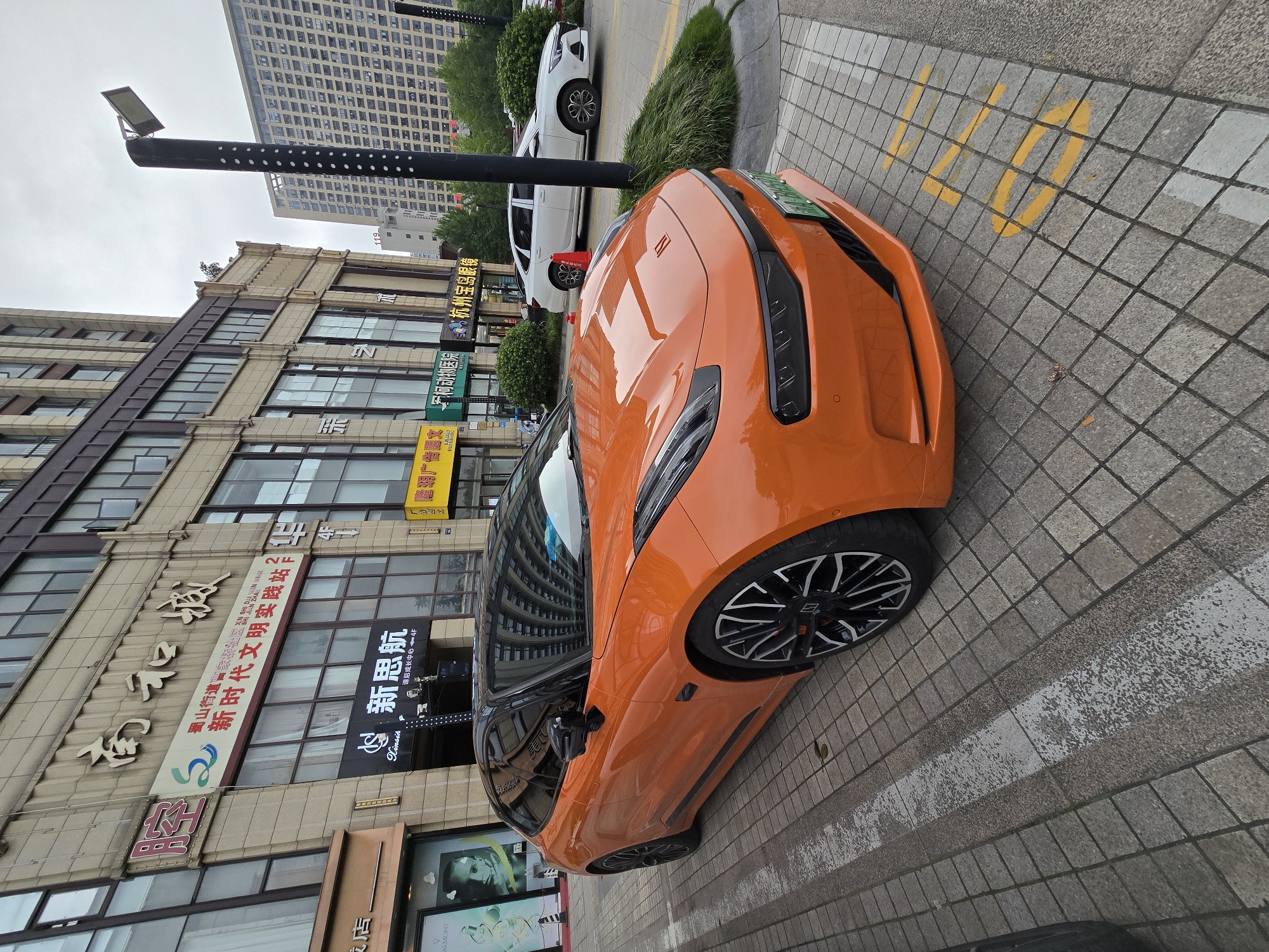 This media file captures an outdoor scene on a city street, likely in Xiaoshan District, China, given the text present. The primary subjects are two cars parked on a tiled pedestrian area. In the foreground, a vibrant orange sports car is prominently displayed, angled towards the viewer. Its sleek design and glossy finish are evident, with its distinctive headlights and alloy wheels clearly visible. Behind it, a white sedan is parked, appearing more conventional in its styling.
The setting is urban, with a multi-story building forming the backdrop. The building features numerous windows and storefronts, some of which display signage with Chinese characters. One banner prominently reads "新时代文明安廠站" and another "新思航." Other visible text includes "Xinsih," "ALMONT," and what appears to be "美" and "课后成长中心-4F."
The ground is paved with square tiles, and yellow painted markings, possibly for parking spaces, are visible. A lamppost with a modern light fixture stands to the left of the orange car. The weather appears to be overcast, contributing to a neutral lighting condition. No people are visible in the frame, and the overall atmosphere is still, suggesting the cars are parked. The image is oriented vertically, giving a sense of looking down on the scene.