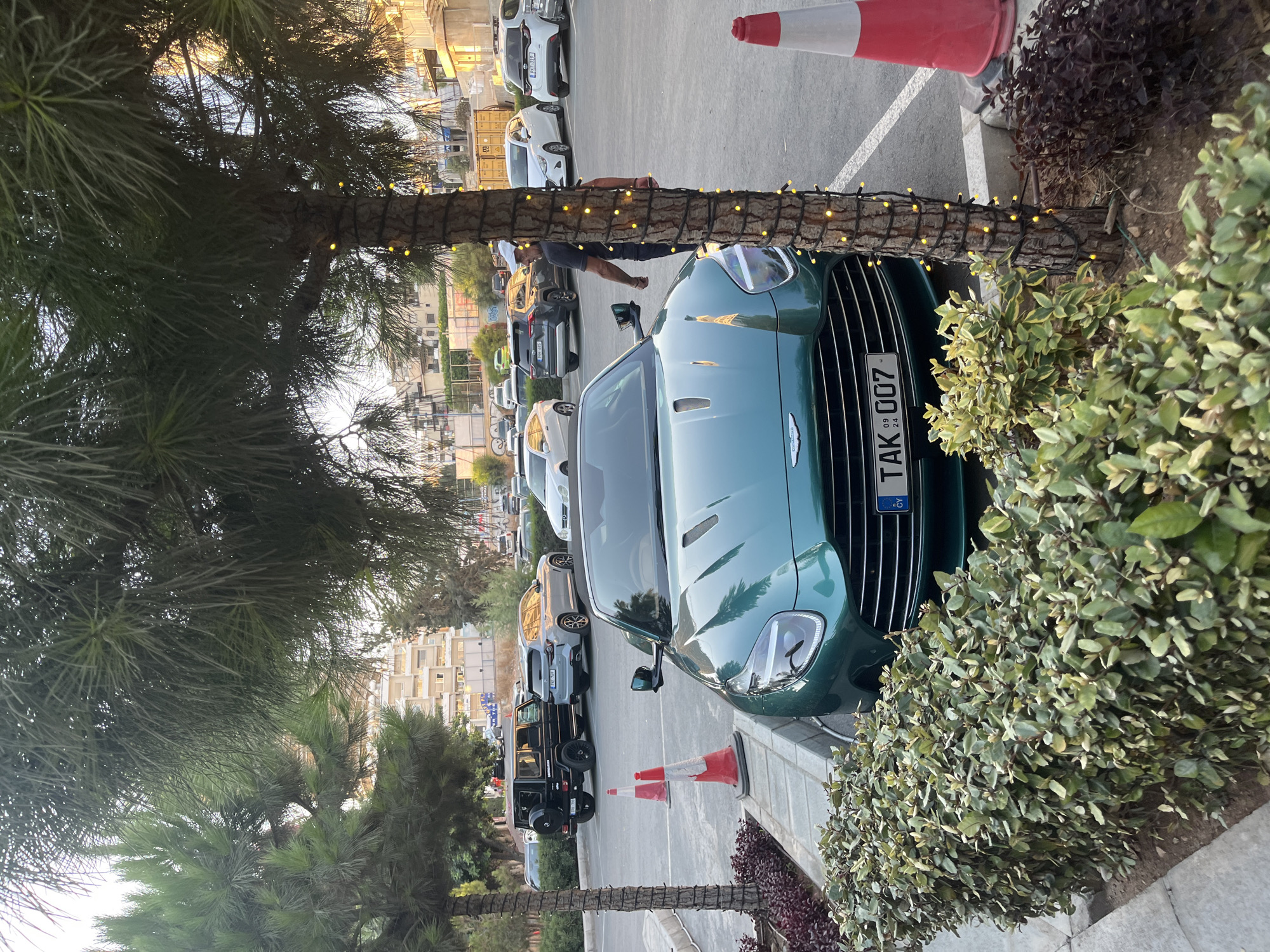 In the city of Agios Tychona Community, Cyprus, a green Aston Martin sports car is parked prominently in the foreground of the image, facing forward. The car features a license plate that reads "TAK 007". Behind the Aston Martin, various other vehicles are parked in a lot, including a black Mercedes-Benz G-Wagon and a grey SUV. A man in a dark t-shirt and white cap is walking away from the viewer towards the right side of the frame. The scene is set in an outdoor parking area with a paved surface. Lush green bushes line the front of the Aston Martin and extend along the left side of the frame. Several palm trees are present, with one in the foreground on the left and another on the right, the latter adorned with yellow string lights. In the background, buildings and more trees are visible. Traffic cones are placed on the pavement. The lighting suggests it is either early morning or late afternoon.
