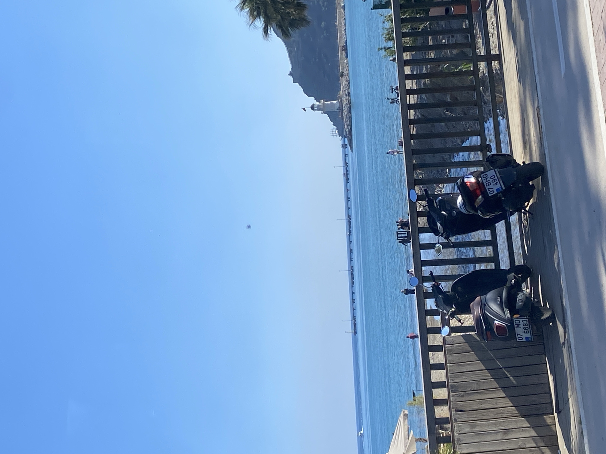 This image captures a vibrant coastal scene in Alanya, Turkey, under a clear, sunny sky. The perspective is slightly elevated, looking out over the sea from what appears to be a promenade or road.
In the immediate foreground, a paved area, likely a roadside or walkway, is visible with a white dividing line. Two dark motorbikes or scooters are parked next to a robust wooden fence or railing. The license plate of the upper scooter clearly displays "07 BHG 490," while the lower scooter's plate reads "07 MD 6883." Both plates bear the "07" prefix, consistent with Antalya Province, where Alanya is located.
Beyond the railing, the serene, turquoise-blue Mediterranean Sea extends towards the horizon. A long, narrow pier or breakwater stretches out into the water, culminating in a distinctive white lighthouse at its far end. In the distance, along the water's edge and on the pier, several small figures of people can be discerned, engaged in activities such as walking or enjoying the water. The distant coastline features a rugged, tree-covered mountain range, and the fronds of a palm tree are visible in the top left corner, adding to the Mediterranean ambiance. The bright, cloudless sky indicates a clear and sunny day, suggesting fair weather and a pleasant atmosphere.