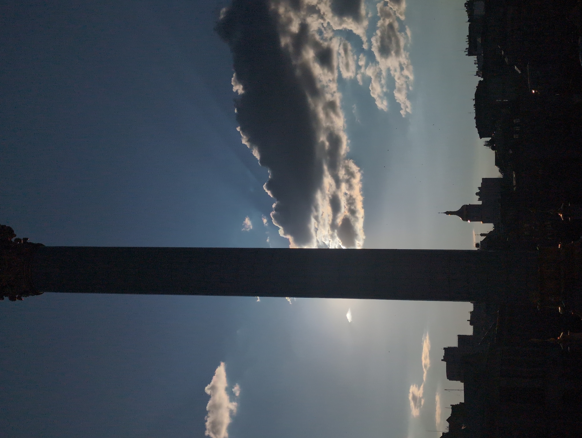A silhouette of a tall column against a dramatic sky is the primary focus of the image. The column, which appears to be part of a monument, stands in the foreground, casting a dark shadow. The sky behind it is a mix of deep blue and lighter shades, with rays of sunlight piercing through some dark, textured clouds. Smaller, brighter clouds are scattered across the sky, illuminated by the sun. In the background, a city skyline is visible in silhouette. The buildings are dark masses, with some rooftops and structures dimly outlined. Notably, a dome-shaped structure with a cross on top is visible on the left side of the skyline, indicative of a church or cathedral. The overall impression is one of a late afternoon or early evening scene, with the sun low on the horizon. The scene is identifiable as Kyiv, Ukraine, likely depicting the Independence Monument or a similar prominent structure.