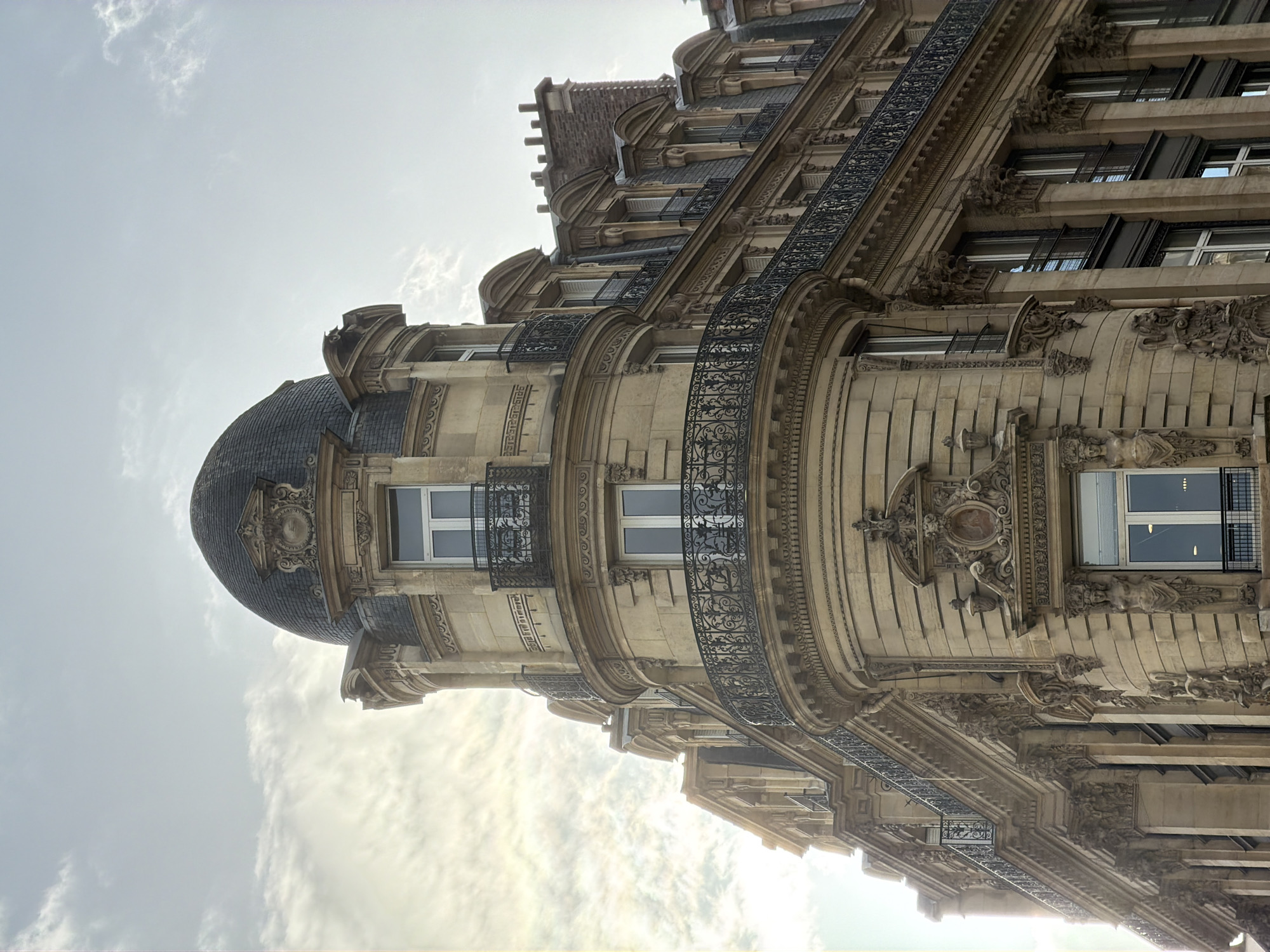 The image depicts the exterior of a classic Parisian building, likely a residential or commercial structure from the late 19th or early 20th century. The architectural style features ornate detailing, including decorative moldings, iron balconies, and a dome-shaped corner turret. The building is constructed of light-colored stone, potentially limestone, which is typical for Parisian architecture. Windows are rectangular with white frames, some with iron bars or railings. The sky above is overcast. The building's location is strongly suggested to be in Paris, France, given the architectural characteristics. No people are visible in the image. The shot is taken from a low angle, looking up at the building's facade.