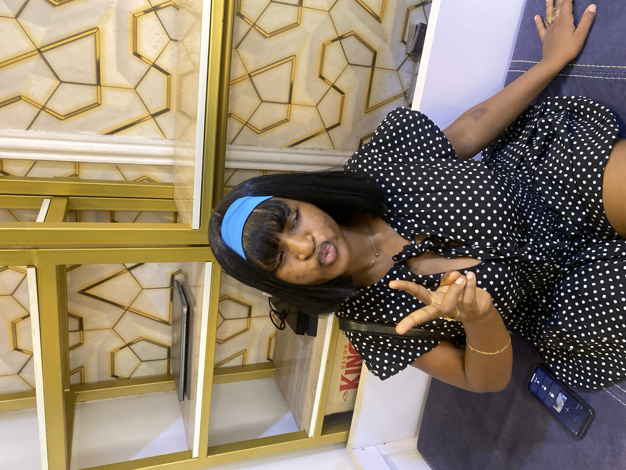 A young Black woman with a blue headband and black bob haircut is sitting on a dark blue bench. She is wearing a black and white polka dot dress with short puff sleeves and a tie at the neckline. Her right hand is raised, displaying a peace sign. She is looking at the camera with her eyes closed and her lips pursed. To her left, on the bench, is a smartphone. Behind her is a gold-trimmed shelf unit with patterned wallpaper. On the shelves, there is a closed laptop and what appears to be a gaming console. The environment is indoors.