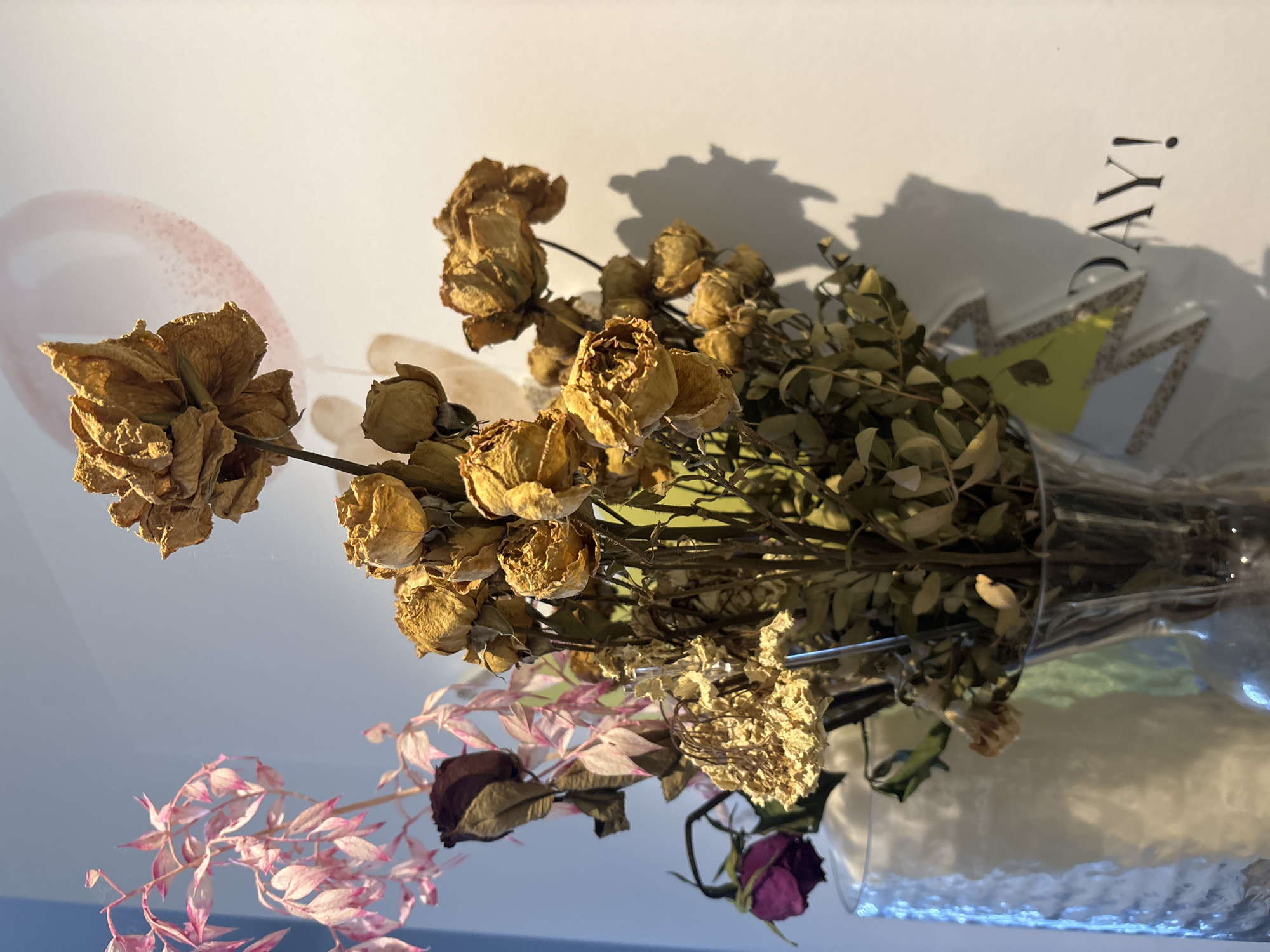 A close-up, eye-level shot captures a bouquet of dried flowers in a glass vase. The bouquet is predominantly composed of small, golden-brown roses, some appearing shriveled and others still retaining their petal structure. Among the roses are delicate, pale pink branches with narrow leaves, and a single, deep purple rosebud. The vase, made of textured glass, sits on a surface. The background is a light-colored wall with a subtle, pale pink circular shape, possibly a painting or decoration, and a shadow cast by the bouquet. To the lower right, a portion of text reads "DAY!" in dark lettering. The scene appears to be indoors. This image was captured in Dubai, United Arab Emirates.