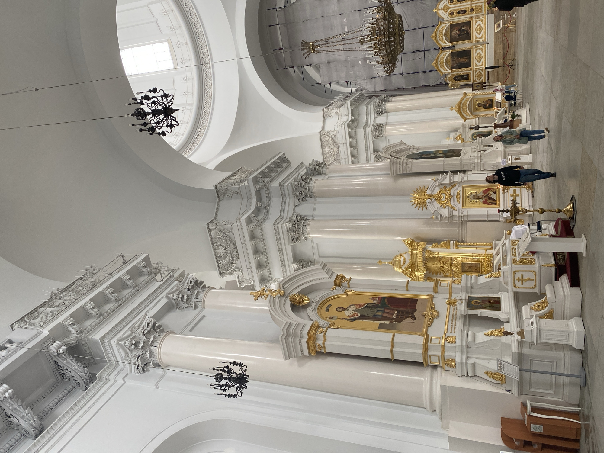 This image shows the interior of Saint Isaac's Cathedral in Saint Petersburg, Russia. The cathedral is a grand and ornate structure with high ceilings, massive columns, and intricate architectural details. The walls and columns are predominantly white, with gold accents on the ornate carvings and iconostasis. Several large chandeliers hang from the ceiling, adding to the opulence of the space.
In the foreground, two people are walking across the marble floor. The person on the left is a young man wearing a black hooded sweatshirt and jeans. The person on the right is a young woman wearing a light blue jacket and jeans. Further in the background, other visitors can be seen milling about, some looking at the icons on the iconostasis. The iconostasis itself is a prominent feature, a large, gilded structure adorned with religious paintings.
The lighting in the cathedral appears to be a mix of natural light coming from high windows and artificial light from the chandeliers. Some areas of the cathedral in the background appear to be under renovation, with scaffolding and protective coverings visible. The overall impression is one of immense scale, spiritual significance, and historical grandeur.