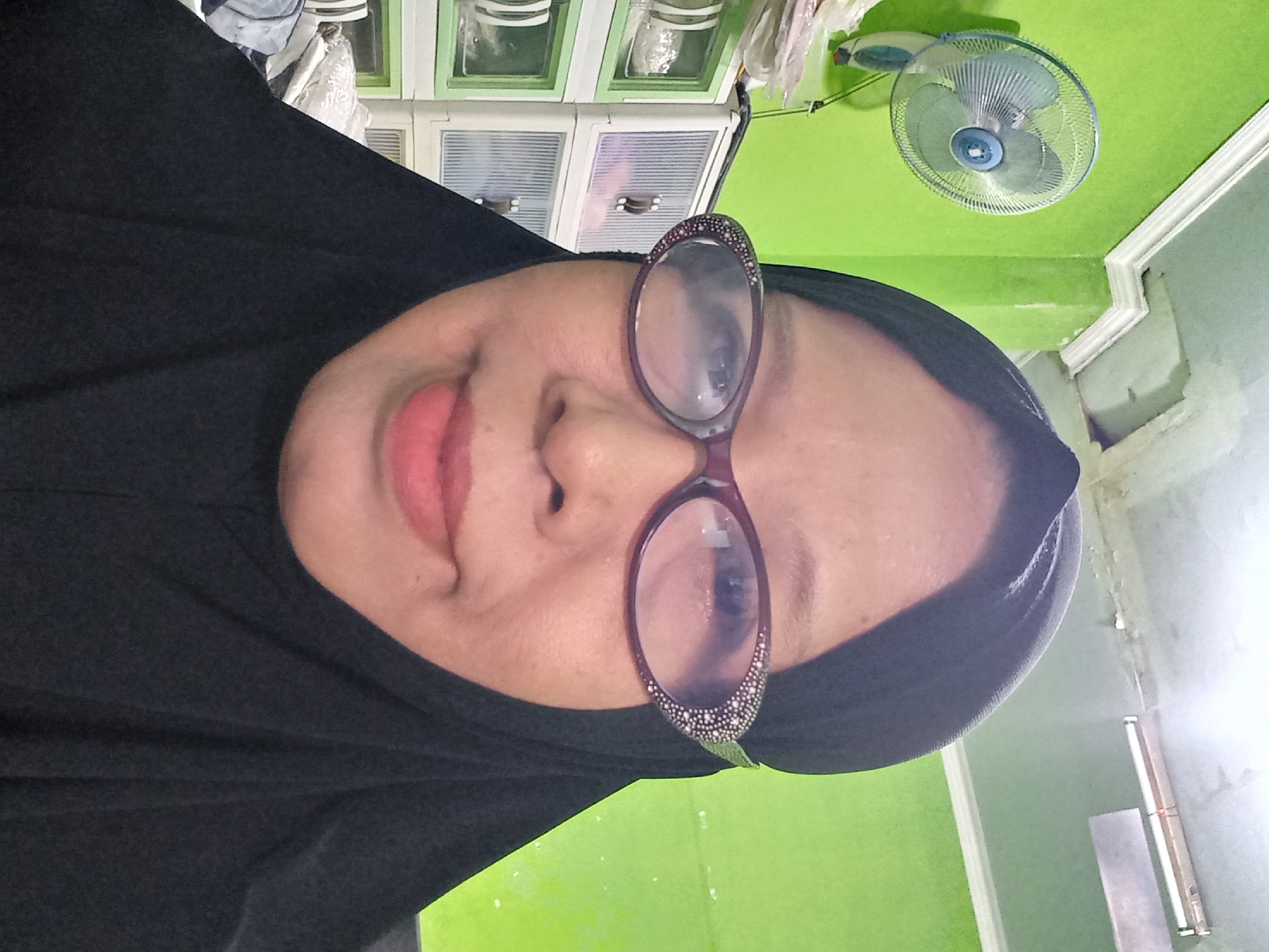 A medium shot shows a woman wearing a black hijab and glasses. She is looking at the camera. The background is a bright green wall and a ceiling with a fluorescent light fixture. To the left of the woman, a white oscillating fan is mounted on the wall. To the right and behind her are clear plastic drawers with white frames. The scene is indoors, suggesting a residential or office setting. The overall impression is a simple, everyday moment captured in a room with vibrant green walls. It is not possible to determine the exact location within Banyumulek, Indonesia from the image alone.