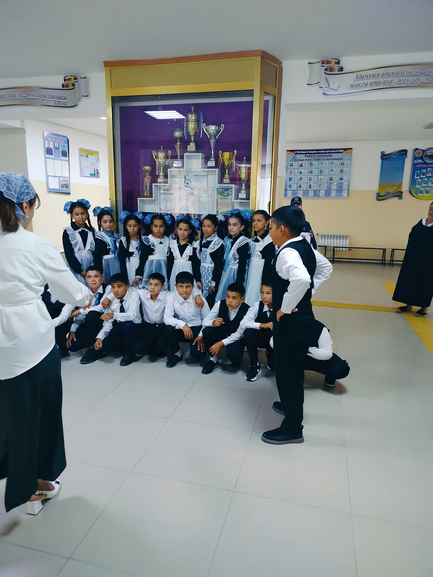 The image captures a group of students, likely posing for a photograph, inside a school hallway in Kölkent, Kazakhstan. The students, dressed in formal school uniforms, are a mix of boys and girls, with the girls wearing blue bows in their hair and the boys in white shirts and dark vests or jackets. Some students are kneeling while others are standing. In the background, a display case filled with trophies and awards is visible, suggesting an achievement-oriented environment. Various posters and informative charts are also mounted on the walls, adding to the institutional setting. A teacher or adult is positioned on the left, facing the students, possibly directing the photo session. The hallway flooring appears to be tiled.