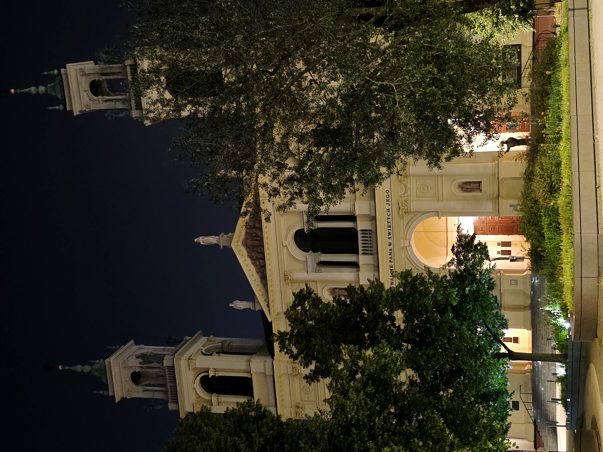 The image depicts the exterior of the Basilica of the Holy Cross in Warsaw, Poland, at night. The imposing church facade is illuminated, highlighting its neoclassical architecture with two prominent bell towers flanking the central structure. Statues adorn the facade, and a large inscription is visible above the main entrance. Dense green trees partially obscure the view of the building. The foreground features a modern plaza with planters and low-lying landscaping, also illuminated by subtle lighting. The overall scene is dark, characteristic of nighttime, with the artificial light sources casting a warm glow on the building and its surroundings. There are no visible people in the image.