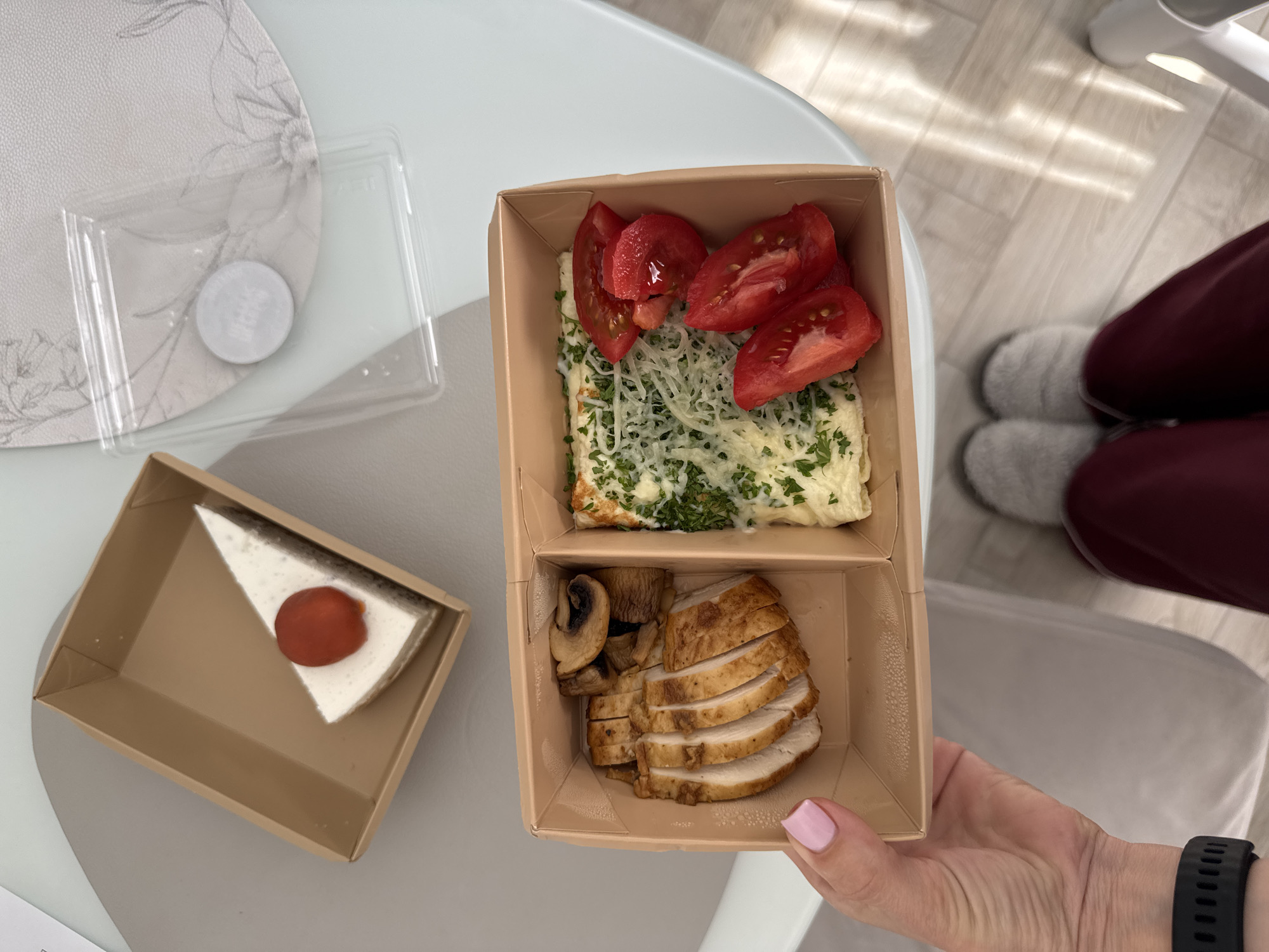 A high-angle vertical shot captures a takeout meal held by a person in an indoor setting, allegedly located in Sochi, Russia.
The primary subject is an open, two-compartment kraft paper takeout container, held by the right hand of an individual in the upper right quadrant of the image. The hand displays neatly manicured pink fingernails. In the upper left quadrant, partially visible, is the person's left lower leg and foot, clad in dark red loose-fitting trousers and light grey, fuzzy slippers. A black fitness tracker or watch is visible on the person's left wrist, just above the hand holding the container.
The left compartment of the main food container holds an egg dish, likely an omelet or scrambled eggs, generously topped with shredded white cheese and finely chopped green herbs, possibly parsley or dill. Four bright red tomato wedges are arranged alongside the egg dish. The right compartment contains several slices of cooked, grilled chicken breast, displaying a browned exterior and white interior, accompanied by two whole sautéed brown button mushrooms.
Below the main meal, on a light grey table surface, a smaller, rectangular kraft paper box is open, revealing a triangular slice of white cheesecake or a similar creamy dessert. It is adorned with a single small, round, vibrant red fruit or decorative candy atop its surface.
To the left of the dessert box, a clear rectangular plastic lid and a small white circular object with faint, illegible text rest on a round white placemat featuring a subtle grey floral pattern. The table surface is a smooth, light grey or bluish-grey. The background features light-toned wooden floorboards illuminated by distinct rectangular sunbeams, indicating natural light streaming into the space. A section of a modern, light grey upholstered chair or sofa armrest is visible in the upper right background.
