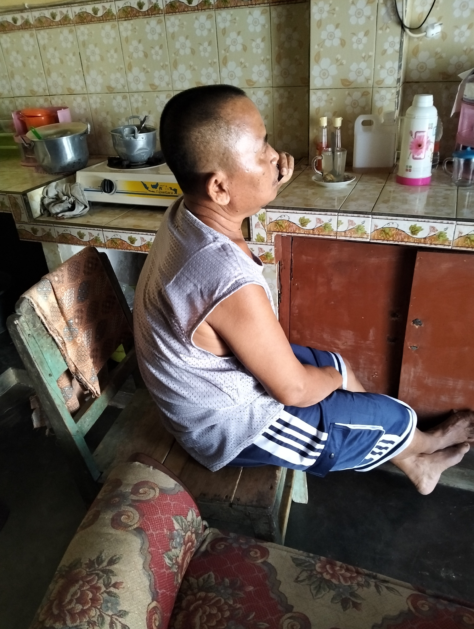 A man sits on a wooden chair in what appears to be a home kitchen or dining area in Sumber Agung, Indonesia. The man is wearing a light-colored, short-sleeved patterned shirt and blue shorts with white stripes. He is facing away from the camera and to the right. In the background, a kitchen counter is visible with various items including pots, a stove, bottles, a thermos, and a plastic jug. The walls are tiled with a floral pattern. A patterned fabric is draped over the wooden chair. Part of a sofa with a floral pattern is in the foreground.