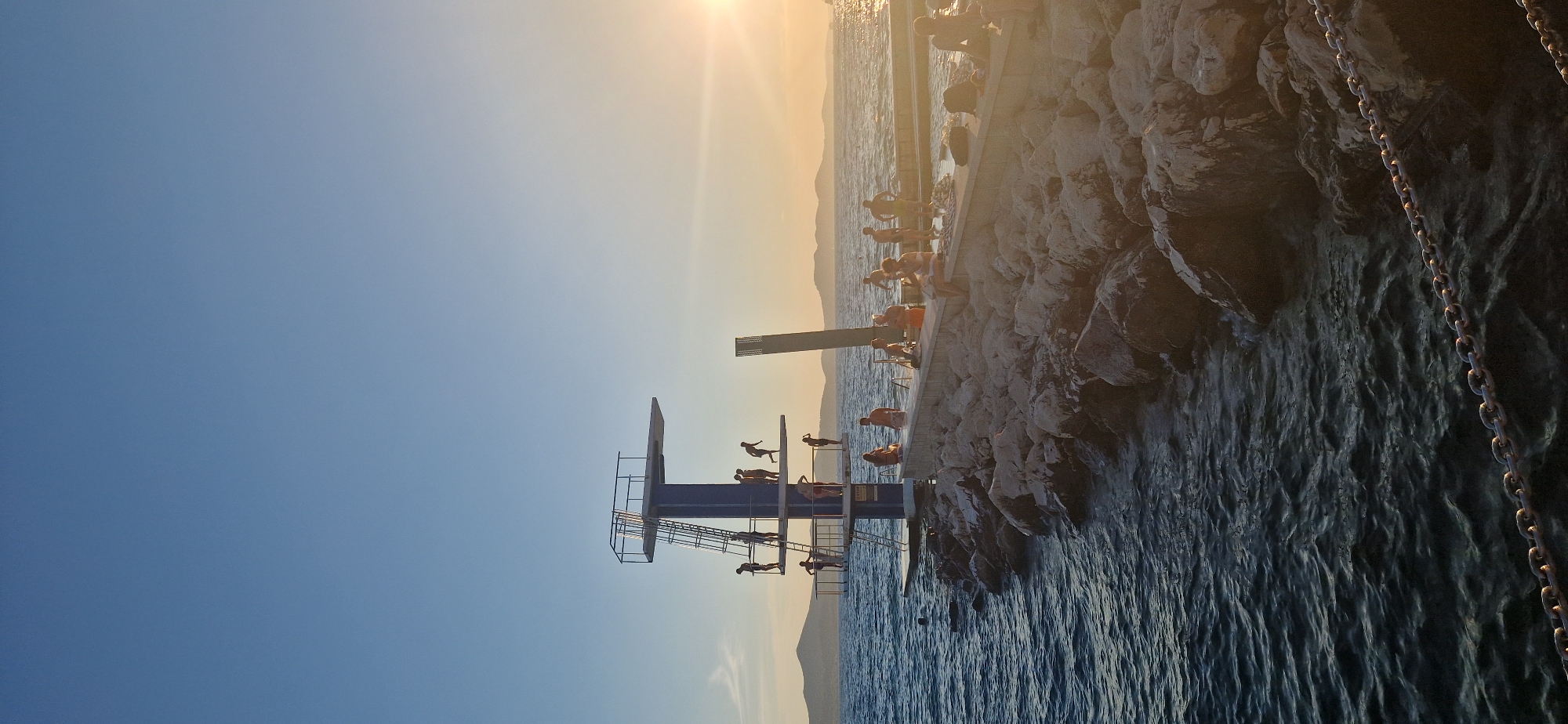 This is a vertical, wide-angle, eye-level photograph captured at sunset, showing a high diving platform situated on a rocky embankment along the sea. The scene is located at the Kolovare Beach in Zadar, Croatia. Several people are visible on and around the diving platform, with some preparing to jump, others on the platform, and some in the water. The sea is dark blue and slightly rippled. The rocky embankment extends into the foreground and is composed of various sizes of light-colored rocks. In the background, the sun is setting over the horizon, casting a warm glow and creating lens flare. Distant islands are silhouetted against the orange and yellow sky. A chain is visible in the lower foreground, running horizontally across the image.