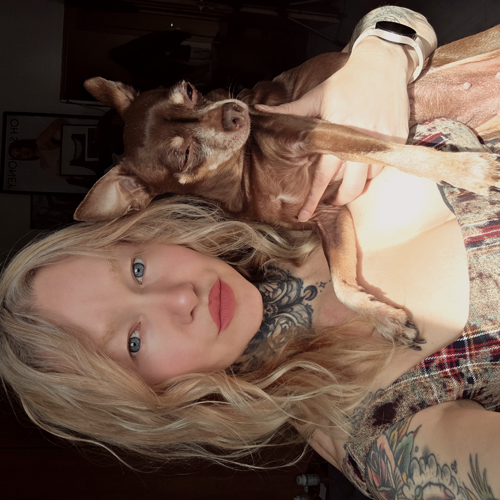 A woman with blonde hair and blue eyes is holding a brown Chihuahua. She has tattoos on her neck and arms. The woman is wearing a plaid shirt. The dog is lying on her chest and appears to be sleeping. The background is dark and out of focus, but a poster on the wall is visible. The location is not specified in the image and there is no way to determine if it is in Saint Petersburg, Russia.