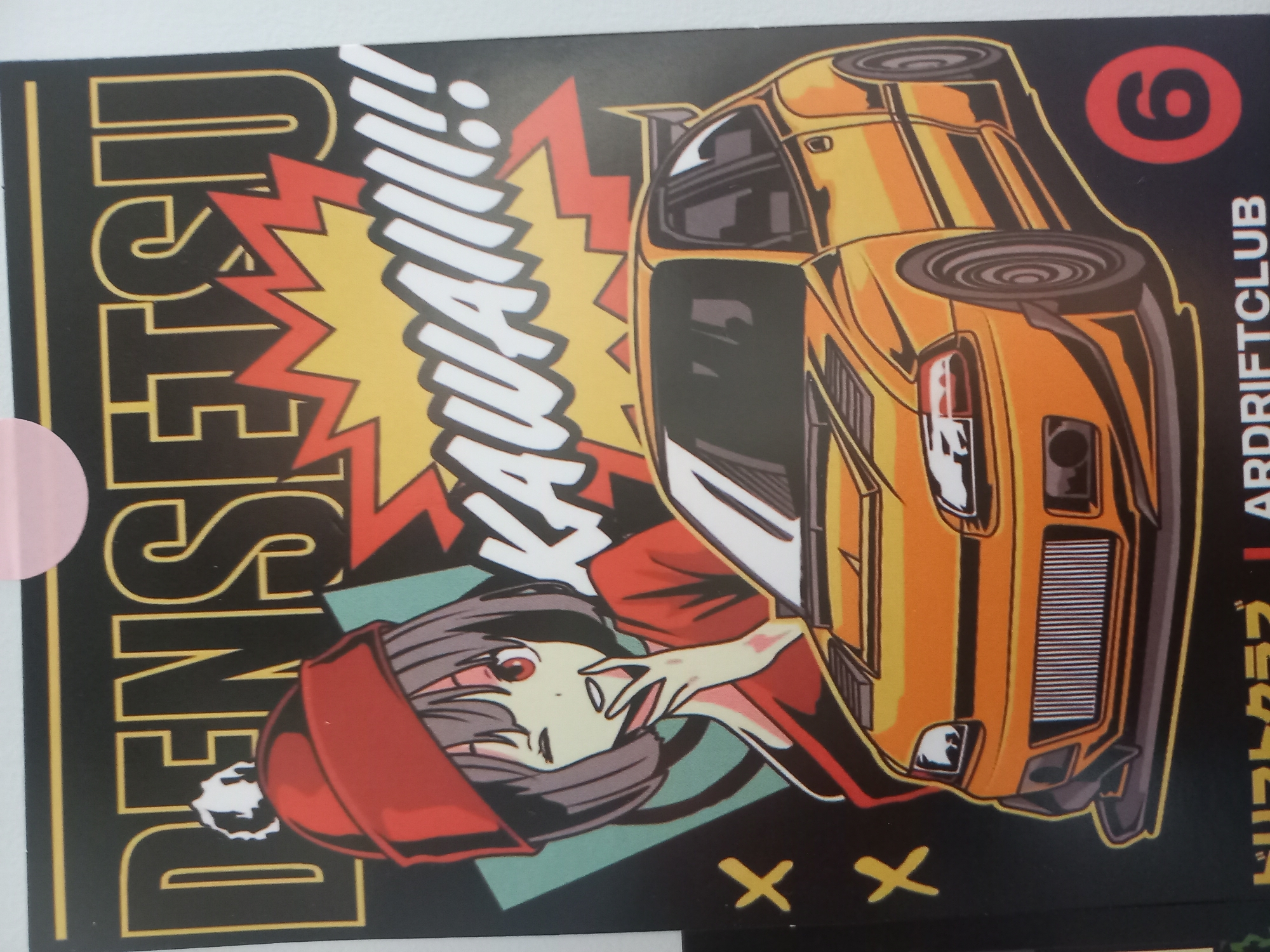 The image is a close-up of a poster featuring an anime-style illustration of a young woman in a red beanie and jacket, winking and touching her face. Behind her, the word "DENSETSU" is written in large, bold yellow letters with a black outline. Below this, the word "KAWAII!!!" is written in white, bubble-like letters with a yellow and red jagged background. In the foreground, a bright orange, modified sports car is depicted with a spoiler and racing decals. The poster has a dark background with some yellow "X" marks on the left side. In the bottom right corner, a red circle with the number "6" inside is visible, and text that appears to be Japanese characters and "ARDRIFTCLUB" is present at the very bottom. The image itself is a photograph of this poster. There is no discernible location or context within the city of Parvomay, Bulgaria.
