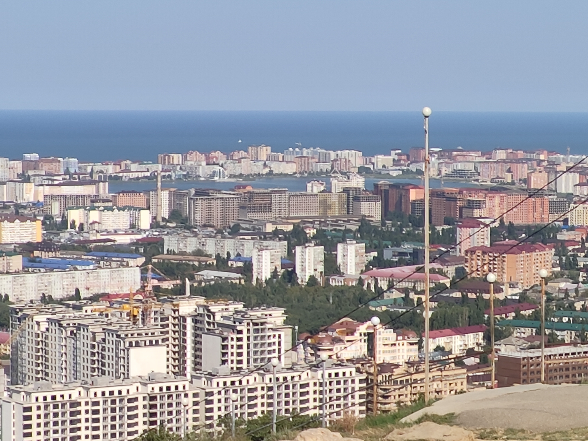 The image shows a panoramic view of Makhachkala, Russia, a coastal city on the Caspian Sea. The foreground features a street lamp with a white globe and some rocky terrain. In the mid-ground, there are several multi-story buildings, some of which appear to be under construction with visible cranes and scaffolding. These buildings are densely packed, creating a cityscape. Further in the background, more buildings of various architectural styles and colors are visible, stretching towards the horizon. The Caspian Sea is prominent in the distance, appearing as a vast expanse of blue water under a clear, light blue sky. The overall scene depicts a developing urban environment with a backdrop of the sea.