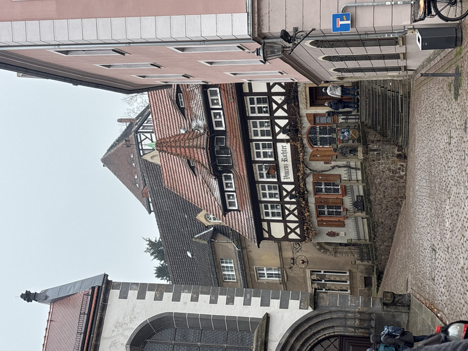 An outdoor street scene in Meissen, Germany, features the Weingut Vincenz Richter building under an overcast sky with traces of snow on rooftops. A cobblestone street slopes downwards in the foreground.
To the left, a large stone building has grey/white ashlar corners, a dark arched entryway, and an arched window above. Its red-tiled roof is topped with a metallic finial.
Center stage is the two-story, half-timbered "Vincenz Richter" building, identified by a sign. Its light grey/beige lower facade has rectangular windows with brown shutters, arched display windows, and decorative dried vines. The upper story displays a darker timber frame with multiple windows. A complex red-tiled roof includes dormers and a prominent conical spire with a green apex. Stone steps, bordered by a rough retaining wall and metal fence, lead to the entrance, where two individuals stand: one in a dark jacket, one in a light grey hooded jacket.
To the right, a multi-story, light pink building with white corner accents has narrow, dark-framed windows. An old-style black street lamp with a security camera is mounted on its corner, with a blue traffic sign (white horizontal bar) below. A trash receptacle and partial bicycle wheel are in the bottom right.
In the background, a light brown building with a dark tiled roof and a circular wrought-iron sign is visible. Further back, another half-timbered building with a brown-tiled gable roof appears. On the far left foreground, two partially obscured individuals are present: one seen from the back with a dark jacket and blue backpack, and another with a visible shoulder and a red object.