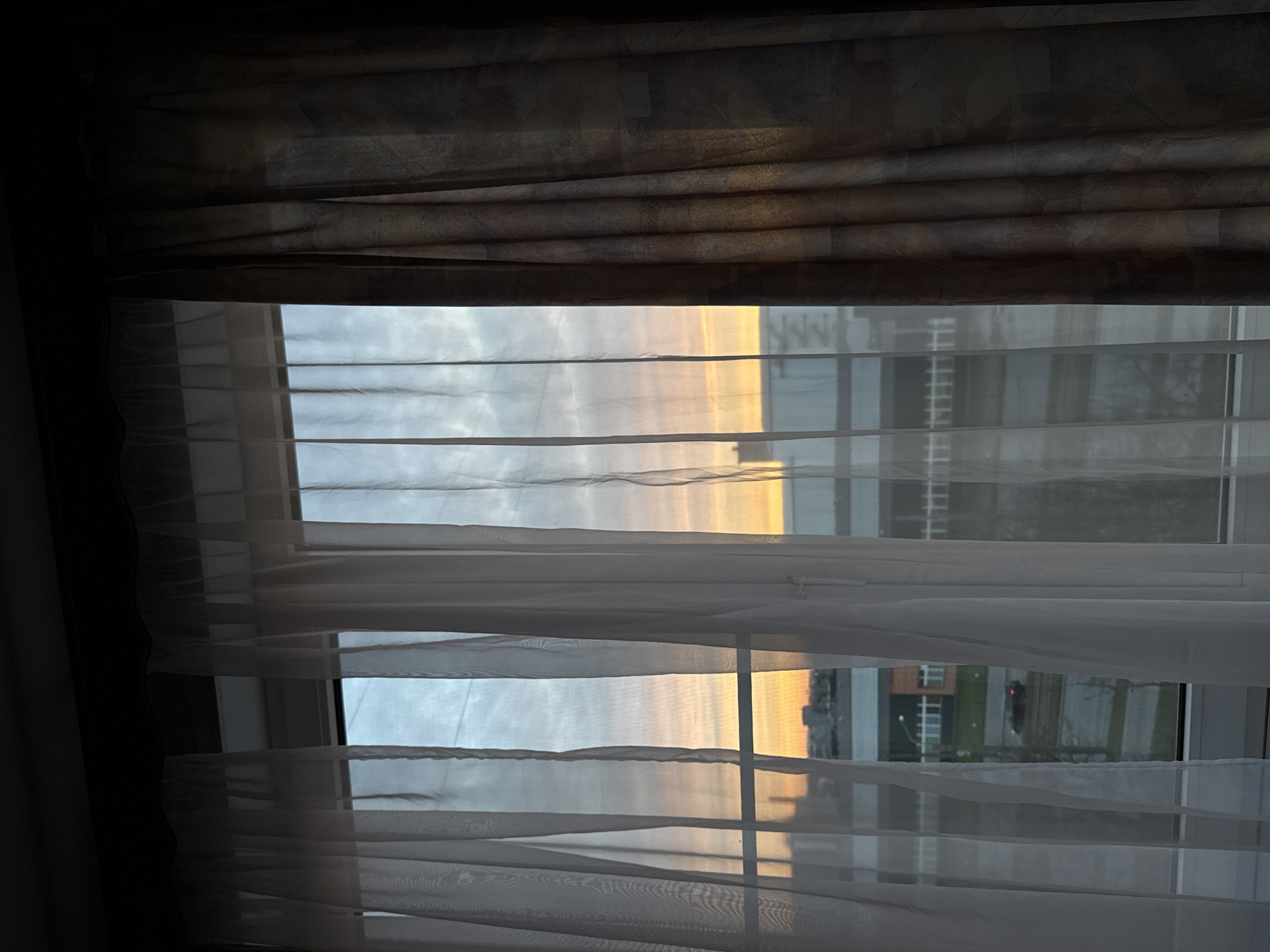 The image shows a view through a window with two layers of curtains. The outer layer is a dark, patterned fabric, partially drawn. The inner layer is a sheer, white fabric, obscuring the view somewhat. Outside the window, a building complex is visible, with a mix of structures and greenery. The sky is visible through the window, with a yellow and grey color gradient, suggestive of a sunrise or sunset. Given the urban features and architectural style, and the prompt's specification, it's consistent with a view in Saint Petersburg, Russia, though lacking the typical prominent landmarks. There is no readily identifiable activity, person, or interaction in the scene.