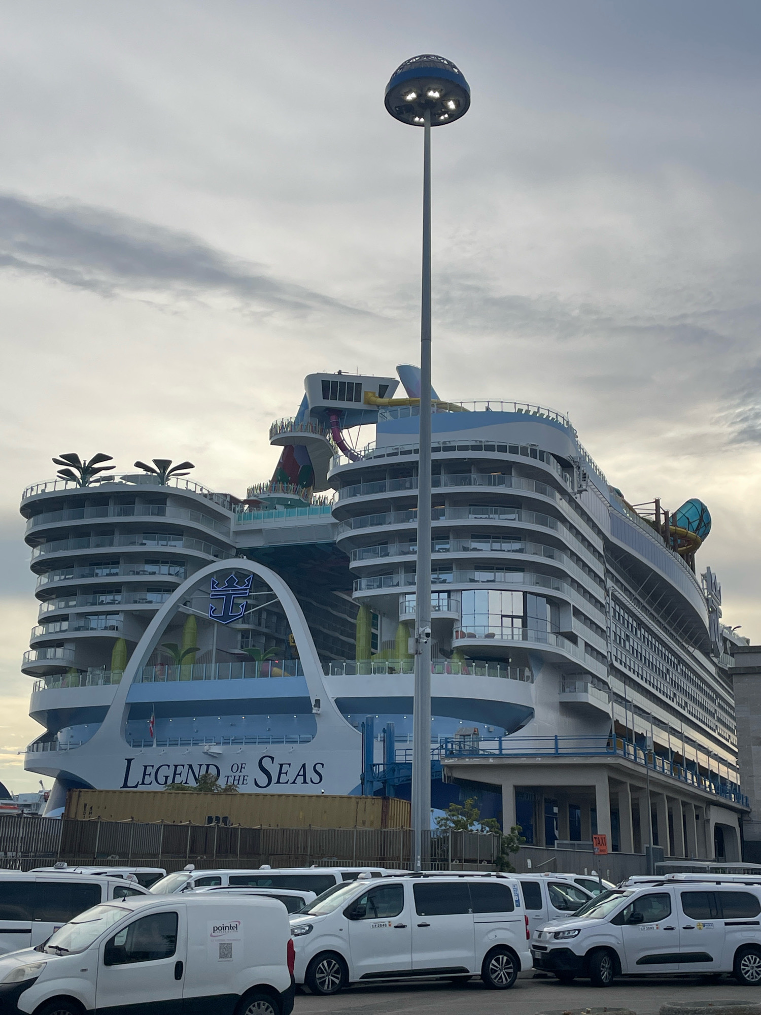 A large white cruise ship, the "Legend of the Seas," is docked in the port of Naples, Italy. The ship has multiple decks with balconies and a water slide on the upper levels. In the foreground, several white vans are parked in a lot, some with markings indicating they are taxis. A tall lamppost with a dome-shaped light fixture stands near the ship. The sky is overcast with clouds.