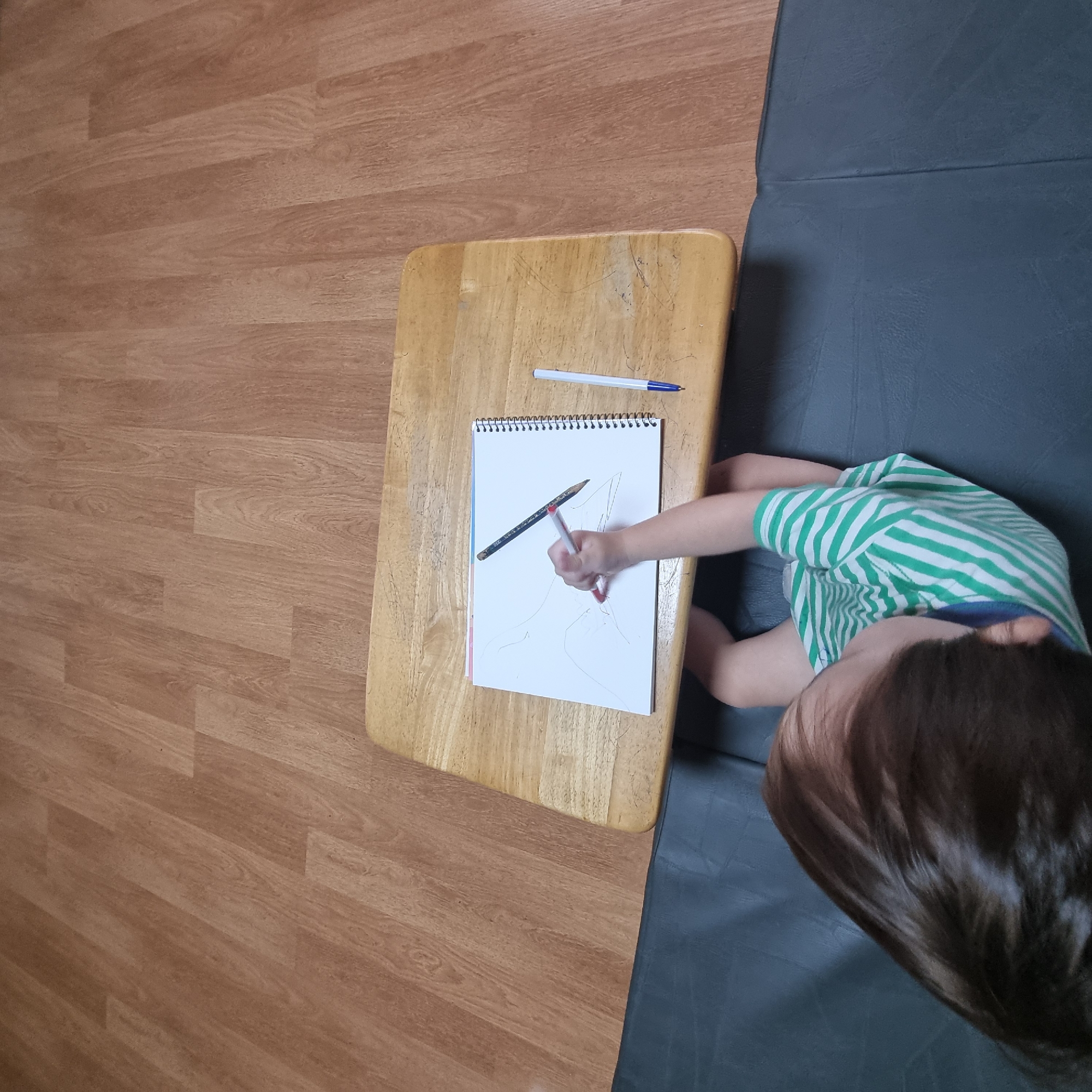 A child wearing a green and white striped t-shirt is sitting on a dark gray mat, drawing on a white sketchbook with a red pen. A black pencil and a white pen with a blue tip are also on the sketchbook. The sketchbook is on a light brown wooden tray table. The child is viewed from behind and from above. The background consists of a light brown wood-paneled floor. The scene is located in Dunnae-myeon, South Korea.