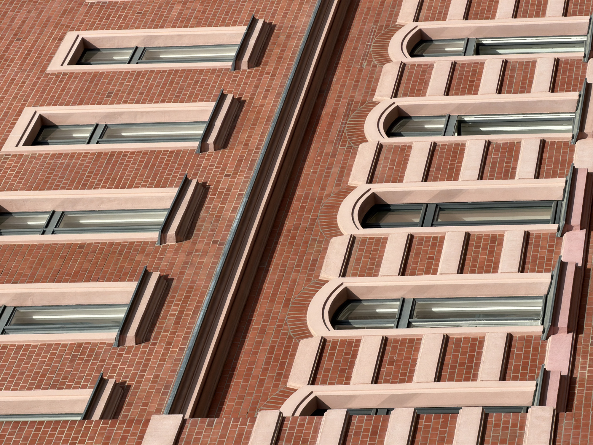 An angled, eye-level close-up shot shows two sides of a red brick building exterior, its distinctive window detailing identifying it as a building located in Berlin, Germany. On the left side of the frame, the windows are horizontal rectangles with pale pink frames that extrude from the brick wall, creating slight overhangs above and below each window. A thin, dark-colored metallic divider separates this side from the one to the right. The windows on the right side of the frame are also horizontal rectangles, but the pale pink frames are more elaborate. They feature vertical pink accents between the windows and arched top portions. All of the windows are reflective, showing some light and shadow inside. The brickwork is consistent across both sides, creating a grid-like texture on the building's surface.