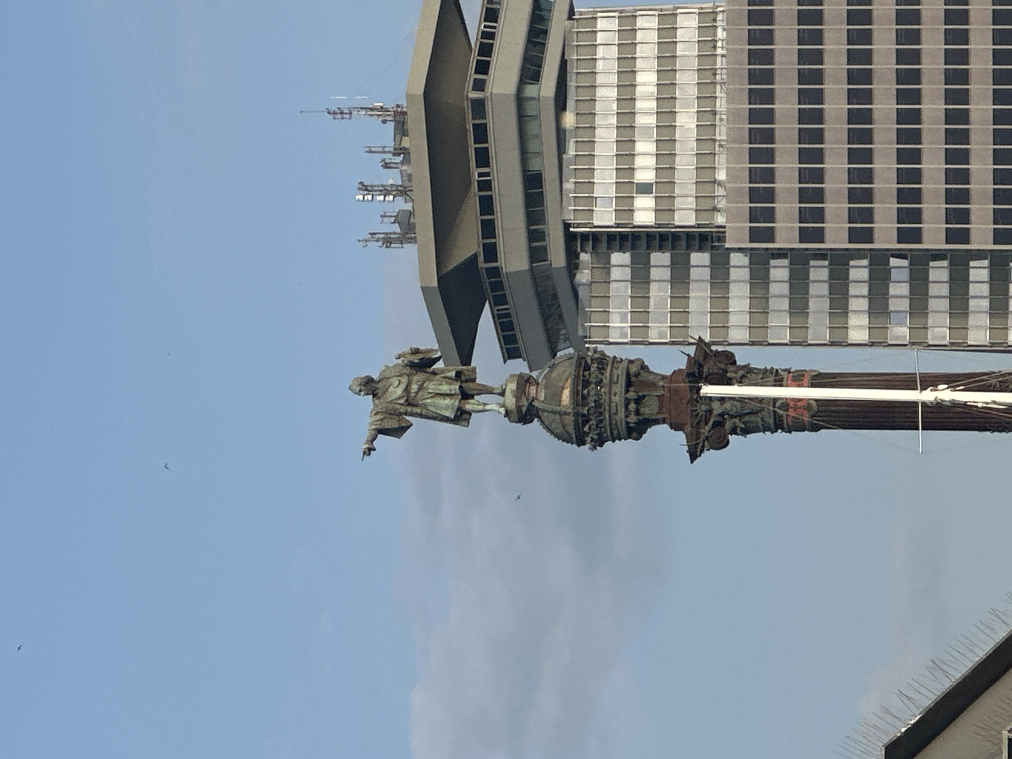 A low-angle, eye-level shot depicts the Christopher Columbus Monument in Barcelona, Spain, juxtaposed with modern buildings and a blue sky. The bronze statue of Columbus, atop an ornate column, points outward. Behind it, tall buildings with grid-patterned facades rise. The sky is pale blue with scattered clouds. In the foreground, a roofline with bird spikes is visible. The monument and buildings dominate the frame, showing a blend of historical and contemporary architecture.