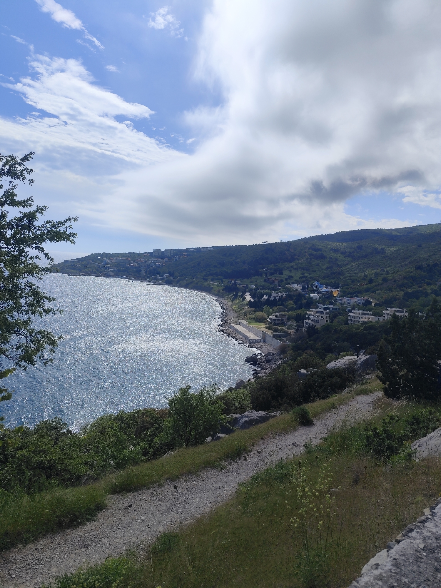 View of the coast and bay in Unknown, Unknown. The coastline curves, and a few buildings are visible on the shore. The sky is partly cloudy. The foreground shows a path with vegetation.