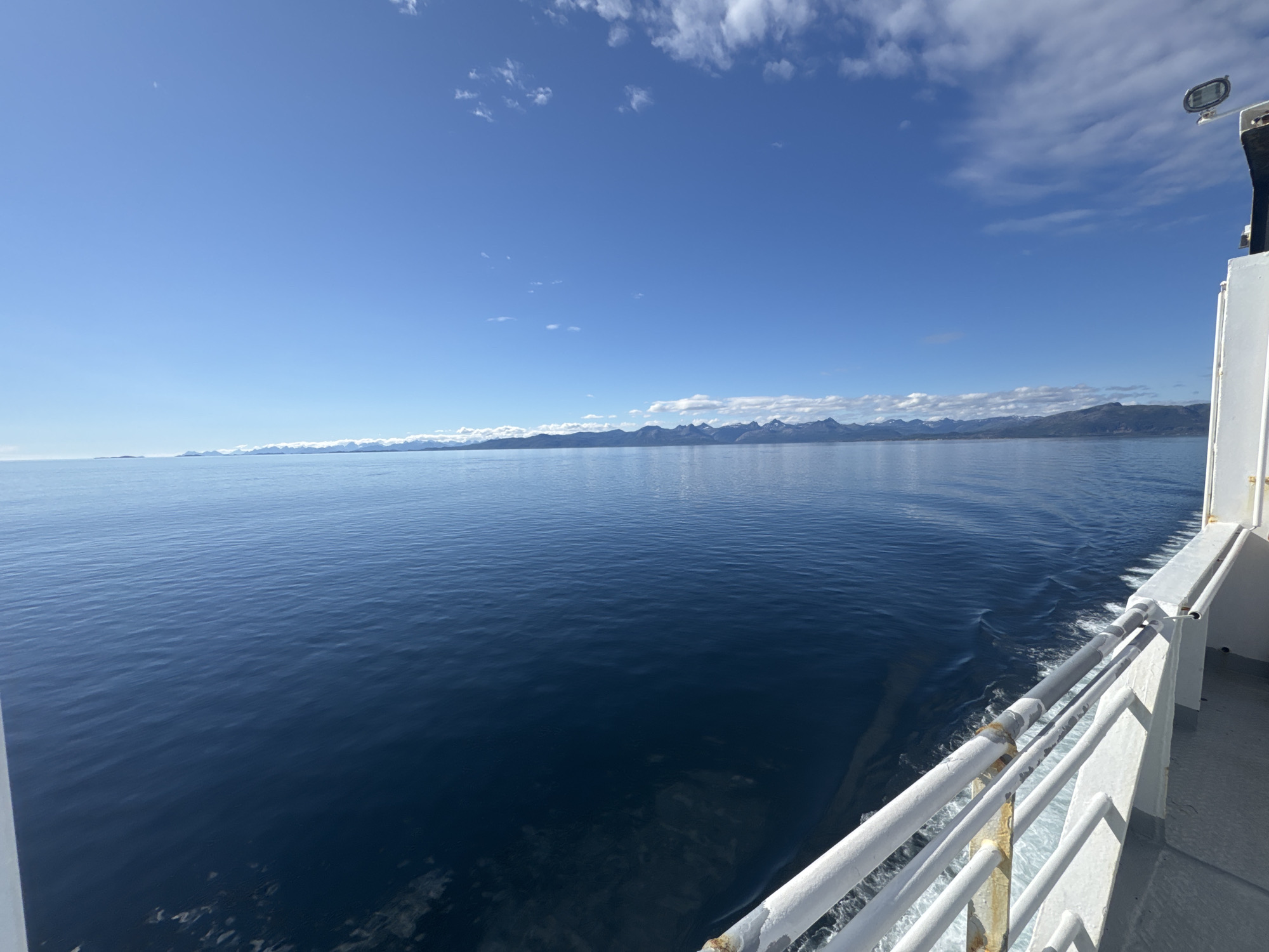 The image shows a wide, calm expanse of deep blue ocean under a bright blue sky dotted with white, wispy clouds. A distant, rugged mountain range with snow-capped peaks lines the horizon, appearing as a dark silhouette against the sky. The perspective is from the deck of a white vessel, with a white railing in the foreground that shows signs of wear, including rust stains on some of the metal supports. White wake trails are visible in the water behind the vessel, indicating it is moving. The sun is shining, suggesting it is daytime, likely midday given the brightness of the sky. The overall mood is serene and vast, with a sense of journey and exploration. The location is hinted to be Norway, which aligns with the mountainous coastal scenery. There is no visible text in the image.
