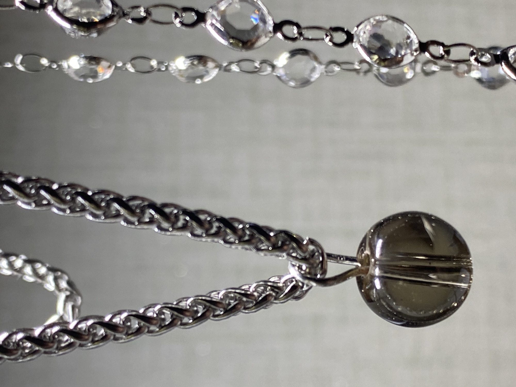The image displays a close-up view of two silver necklaces. The necklace on the left features a thick, woven chain with a dark, round bead pendant. The necklace on the right is more delicate, composed of a series of interconnected oval links, each adorned with a clear, faceted gemstone. The background is a neutral, textured gray surface. There is no discernible information or visual cues within the image to suggest a location in Kushchovskaya, Russia, or any specific environmental context beyond the studio-like setting. No people or actions are depicted.