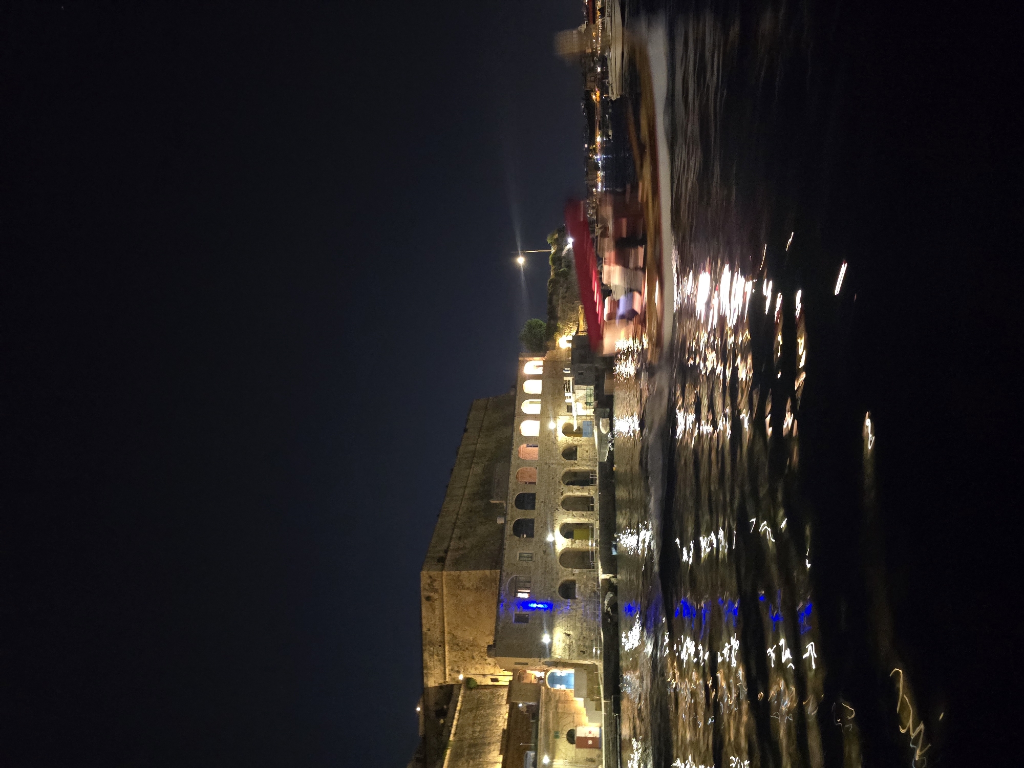 An outdoor, eye-level, long exposure, night shot depicts the historic waterfront of Valletta, Malta, with a fast-moving boat in the foreground. The water, which takes up the bottom half of the frame, is dark and textured, with streaks of reflected light from the buildings and the boat creating a dynamic, abstract pattern. A medium-sized boat with a red canopy and several figures seated within it is captured in motion, appearing blurred due to the long exposure. The boat is heading from left to right across the frame.
The upper half of the image shows a stone building complex along the waterfront. This structure is illuminated by artificial lights, highlighting its arches and windows. A large, fortified wall with a textured surface, characteristic of Valletta's historic defenses, rises behind the waterfront buildings. The sky above is dark and clear, with a single streetlamp casting a beam of light. The overall impression is one of a lively night scene on the water, set against the backdrop of ancient fortifications.