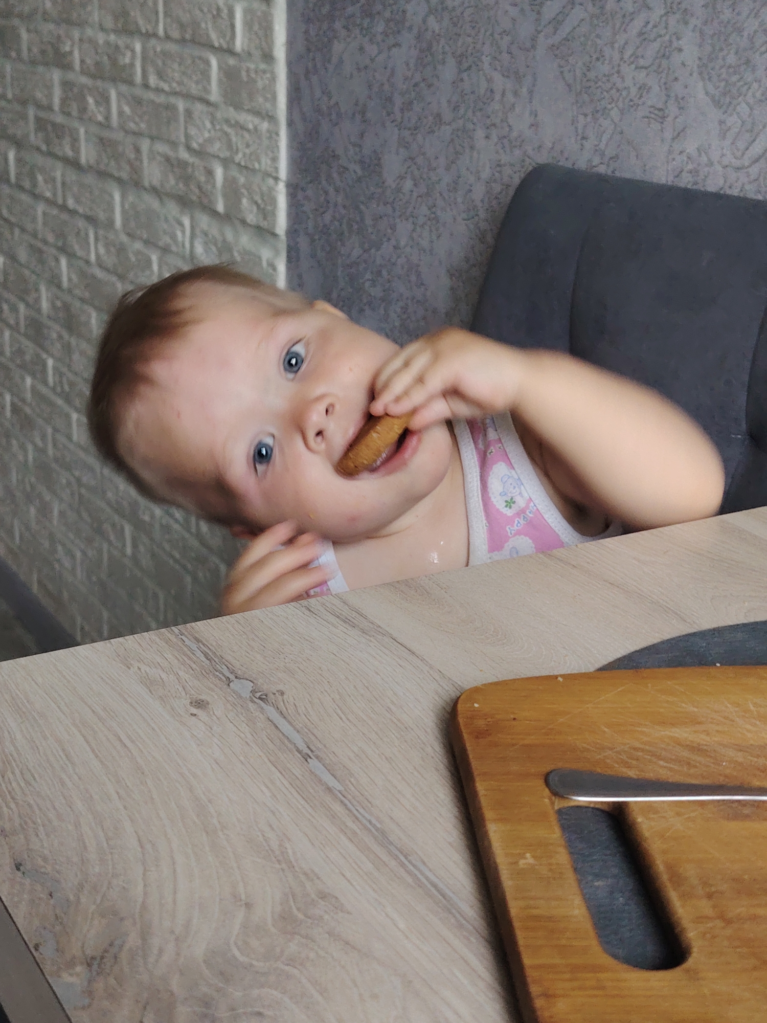 A baby with blue eyes and light brown hair is sitting at a wooden table in what appears to be a domestic setting. The baby is in the process of eating a circular brown object, possibly a cookie or biscuit, and is looking towards the viewer. The baby is wearing a white and pink tank top with a cartoon design. The table surface is light-colored with wood grain patterns. Partially visible on the right side of the table is a brown wooden cutting board with a silver handle.
Behind the baby is a dark gray chair and the background consists of a gray brick wall on the left and a gray patterned wall on the right. Based on the origin of the image, the location is Chaikovsky, Russia.