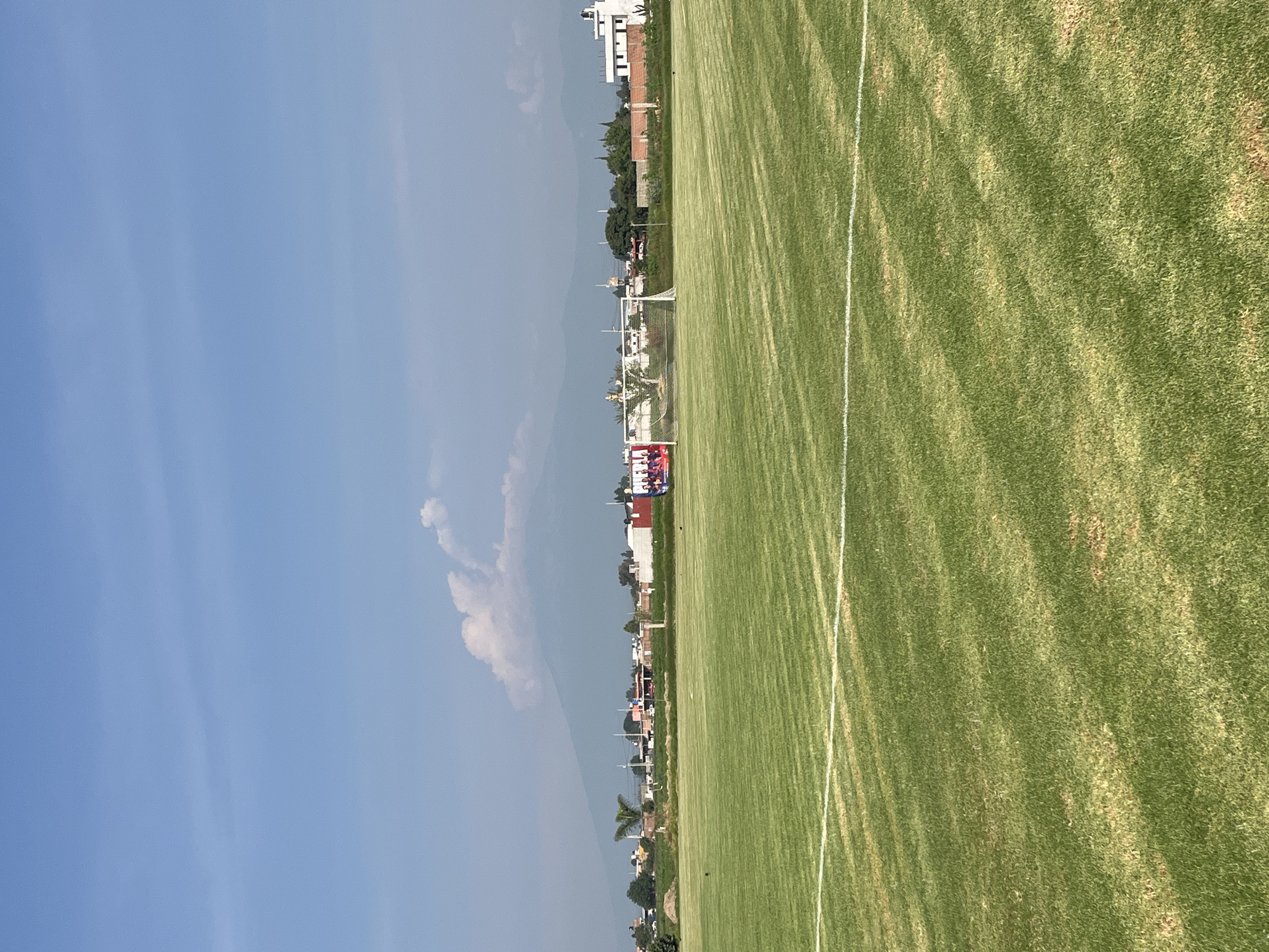 The image captures a partial view of a soccer field in Unknown, Unknown on a sunny day. The grass is green, and marked with white lines. Buildings and trees are visible in the background. The sky is blue with a few scattered clouds.