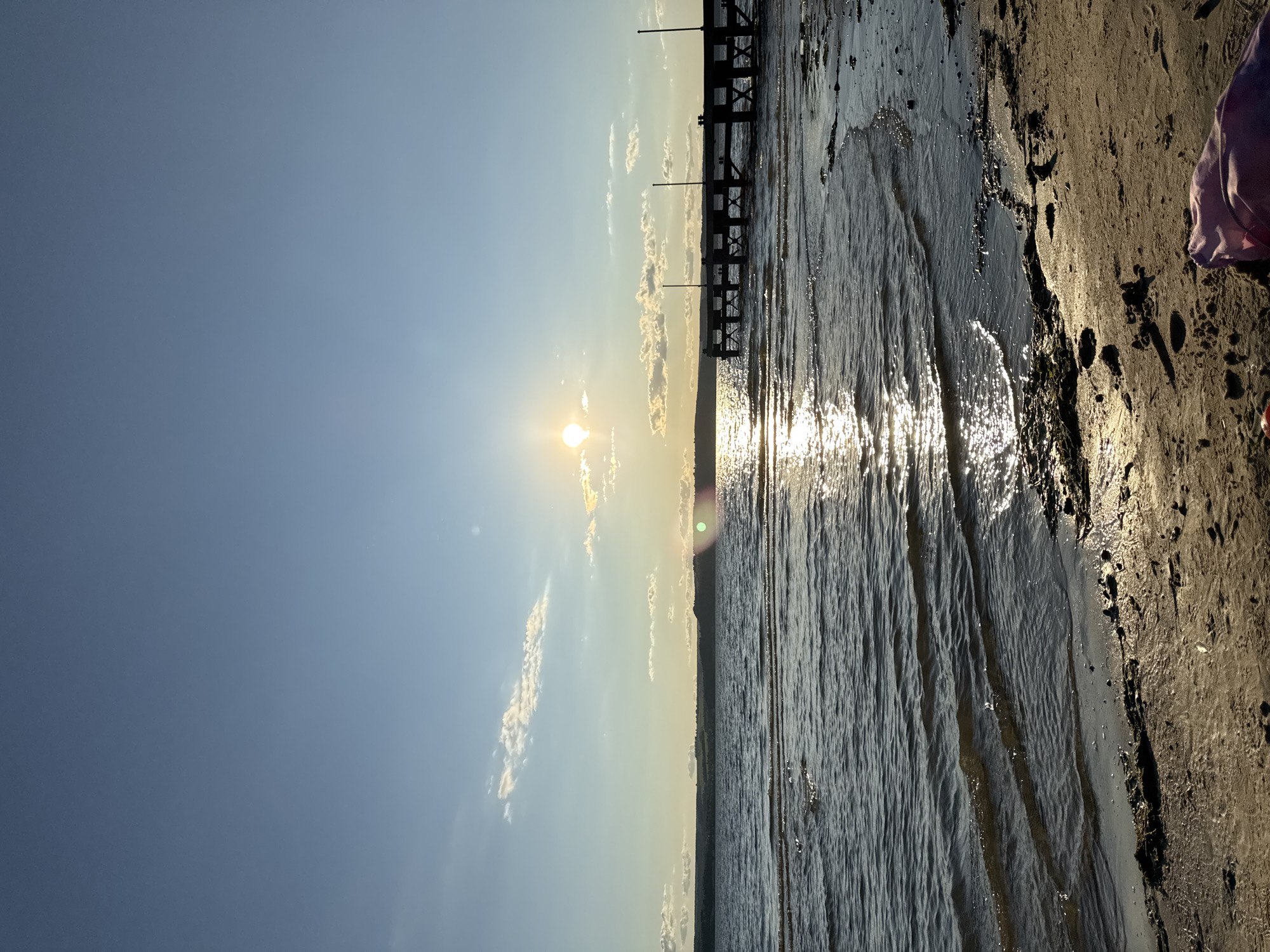 The image shows a sunset over the water in Matyushino, Russia. The sun is low on the horizon, casting a warm glow across the sky and reflecting on the water. There are a few clouds in the sky, and a distant shoreline with rolling hills can be seen. In the foreground, a wooden pier extends into the water. The beach is sandy and wet from the incoming tide. The overall scene is peaceful and serene.