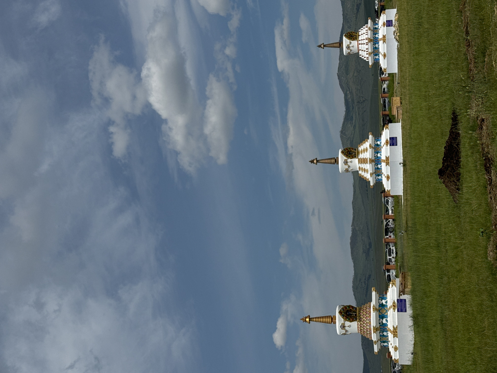 Three white stupas with gold spires stand in a grassy field under a blue sky with scattered clouds. The stupas are ornate, featuring decorative bands of blue and gold, and small figures. In the background, a line of parked white vehicles is visible behind a fence, with rolling green hills beyond. The ground in the foreground is covered in tall green grass, with a pile of dark soil to the right. This scene is located in Orongoi, Russia.