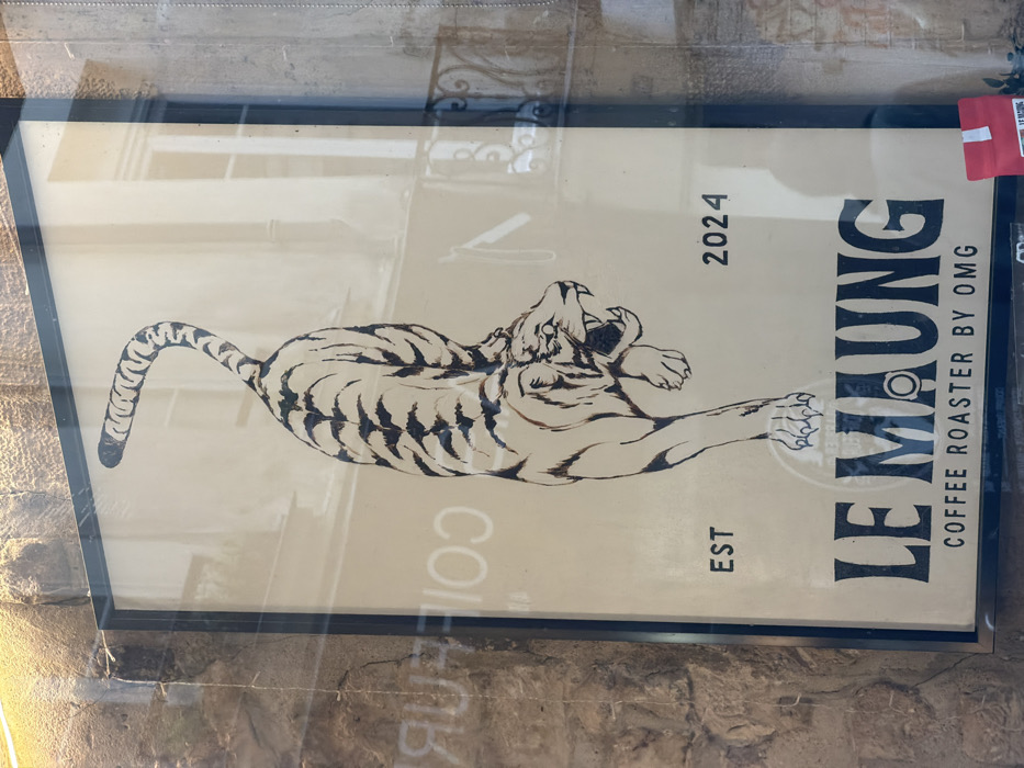 A rectangular, dark-framed sign is observed through a reflective glass panel. The sign features a stylized illustration of a tiger, rendered with dark stripes on a light, beige background, depicted in a crouched, predatory stance with an open mouth and visible fangs. Its tail curves upward and left. Text on the sign reads "EST" to the lower left of the tiger, "2024" to its lower right, and "LE MAUNG" in large, dark, serif typeface centered below the tiger. Beneath this, "COFFEE ROASTER BY OMG" is written in smaller, dark, sans-serif text.
The reflective glass surface shows the reversed word "COIFFEUR" on the left and architectural details of a building across the street, including multiple light-framed windows and a segment of a wrought-iron balcony, consistent with typical Parisian urban structures. The immediate left border of the sign is a rough, light-brown stone wall. A small, red retail package, partially visible at the bottom right, bears the "LE MAUNG" brand. The scene is located on a street in Paris, France.
