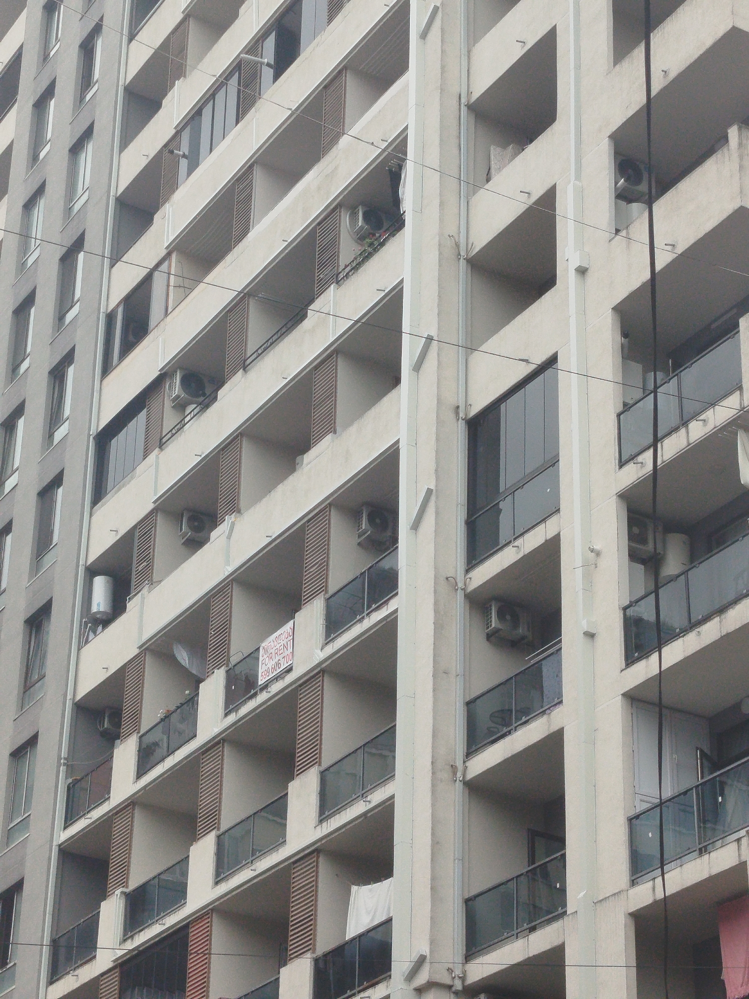 The image shows a close-up, angled view of a multi-story apartment building with a light-colored facade and balconies. The building appears to be located in Batumi, Georgia, as indicated by a "FOR RENT" sign in Georgian script on one of the balconies. The sign also includes an English translation and a phone number. The balconies are furnished with glass railings and some have visible air conditioning units mounted on the walls. Laundry is also visible hanging on some balconies. The building has numerous windows, some of which are tinted. Electrical wires are visible strung across the building's facade. The overall impression is of a residential building in an urban environment.