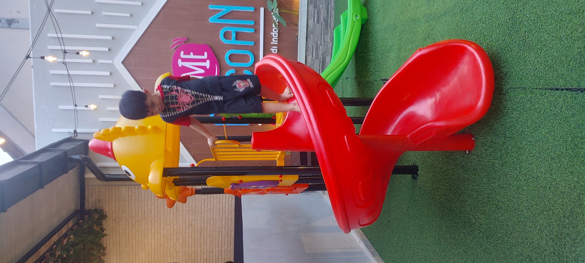A young East Asian boy, appearing to be around 4-6 years old, is standing on the top edge of a red spiral slide in an outdoor playground. He is wearing a black t-shirt with a red graphic on the chest and red sleeves, and black shorts with a similar graphic. His feet are bare. The playground equipment is colorful, with a large yellow structure resembling a fish at the top of the slide, featuring orange accents and a red fin. A yellow railing is visible behind the boy. To the right of the red slide, a green slide is partially visible. The ground beneath the playground is covered in green artificial turf. In the background, a building with a light brown wall and white vertical accents is visible. The building has a sign with pink and light blue lettering, reading "ME" and "COAN". There are strings of lights hanging above the playground. The setting appears to be a public or commercial space, possibly a cafe or restaurant with a play area, and it is located in the city of Jayapura, Indonesia.