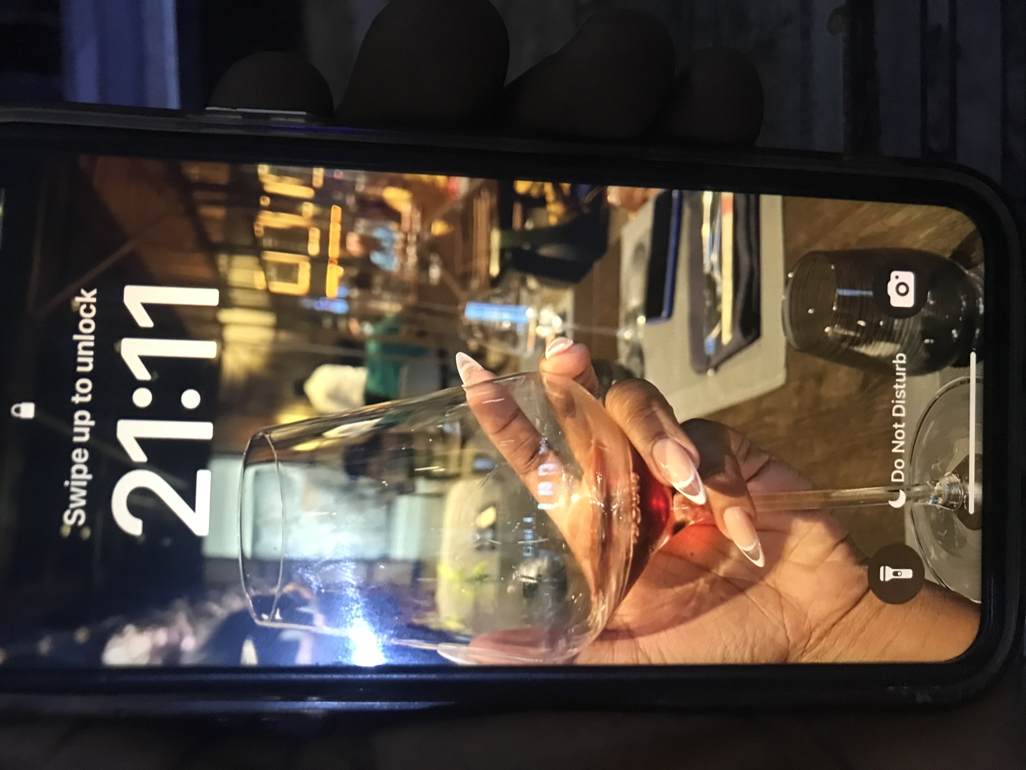 The image shows a black smartphone displaying the lock screen, which reads "Swipe up to unlock" and the time "21:11". The background image on the lock screen depicts a hand holding a wine glass containing a red liquid. The hand holding the glass is dark-skinned with manicured nails. The background of the image is a blurred interior scene with lights and dining ware. There is no information in the image indicating that this scene is located in Kabong, Nigeria.