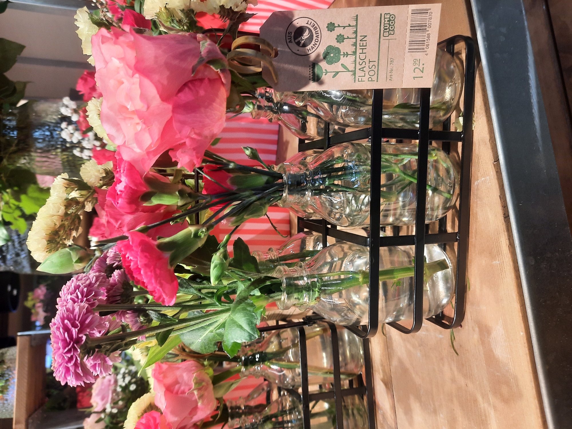 A close-up, eye-level, horizontal shot shows a display of small bouquets of flowers in clear glass bottles. The bottles are arranged in black metal racks on a wooden surface. The flowers are a mix of pink roses, pink carnations, and pale yellow flowers with some delicate white flowers mixed in. The flowers are fresh and vibrant. A brown paper tag is attached to one of the racks, featuring a circular logo with the text "AUS HANDARBEIT" and a drawing of flowers. Below the logo, the text "FLASCHEN POST" is visible, along with "Art-Nr.: 787", a price of "12.99", and a barcode. The background is out of focus, showing more flowers and a striped pink and white surface. The overall scene suggests a florist shop or a market stall, likely in Rosenheim, Germany, given the German text on the tag. There are no people visible in the image.