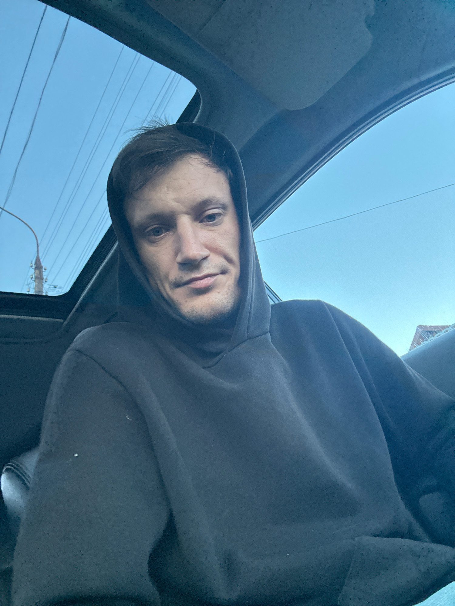 A man wearing a dark hooded sweatshirt is inside a car, looking at the camera. The sunroof of the car is open, revealing a light blue sky with some power lines visible. The man's face is partially obscured by the hood. The background shows a blurry cityscape, suggesting an urban or suburban setting. The image appears to be a selfie taken by the man in the car. The location of Makiv, Ukraine, is not discernible from the visual information present in the image.