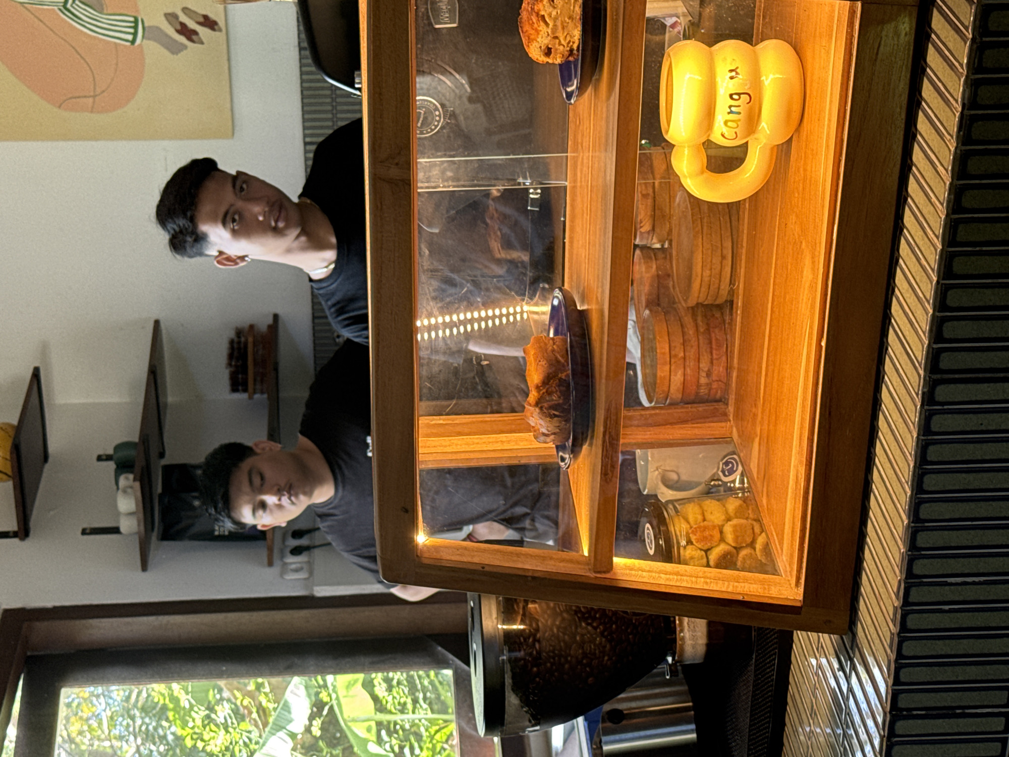 The image depicts the interior of a cafe, likely located in Mengwi, Indonesia, as suggested by the presence of a yellow mug with "Cang" written on it. Two young men, appearing to be employees or patrons, are visible. One man in the foreground, wearing a black t-shirt, stands behind a wooden display case illuminated by warm interior lights. He is looking towards the viewer with a neutral expression. The display case contains various food items, including pastries on blue plates and jars of cookies. Behind him, another young man in a black t-shirt is partially visible, looking to his left. To the left of the scene, a large coffee grinder filled with beans is positioned near a window that offers a view of lush greenery. The cafe's decor includes shelves with decorative items and a framed artwork on the wall. The counter in front of the display case is tiled with a mosaic of small, rectangular tiles in shades of green and black.