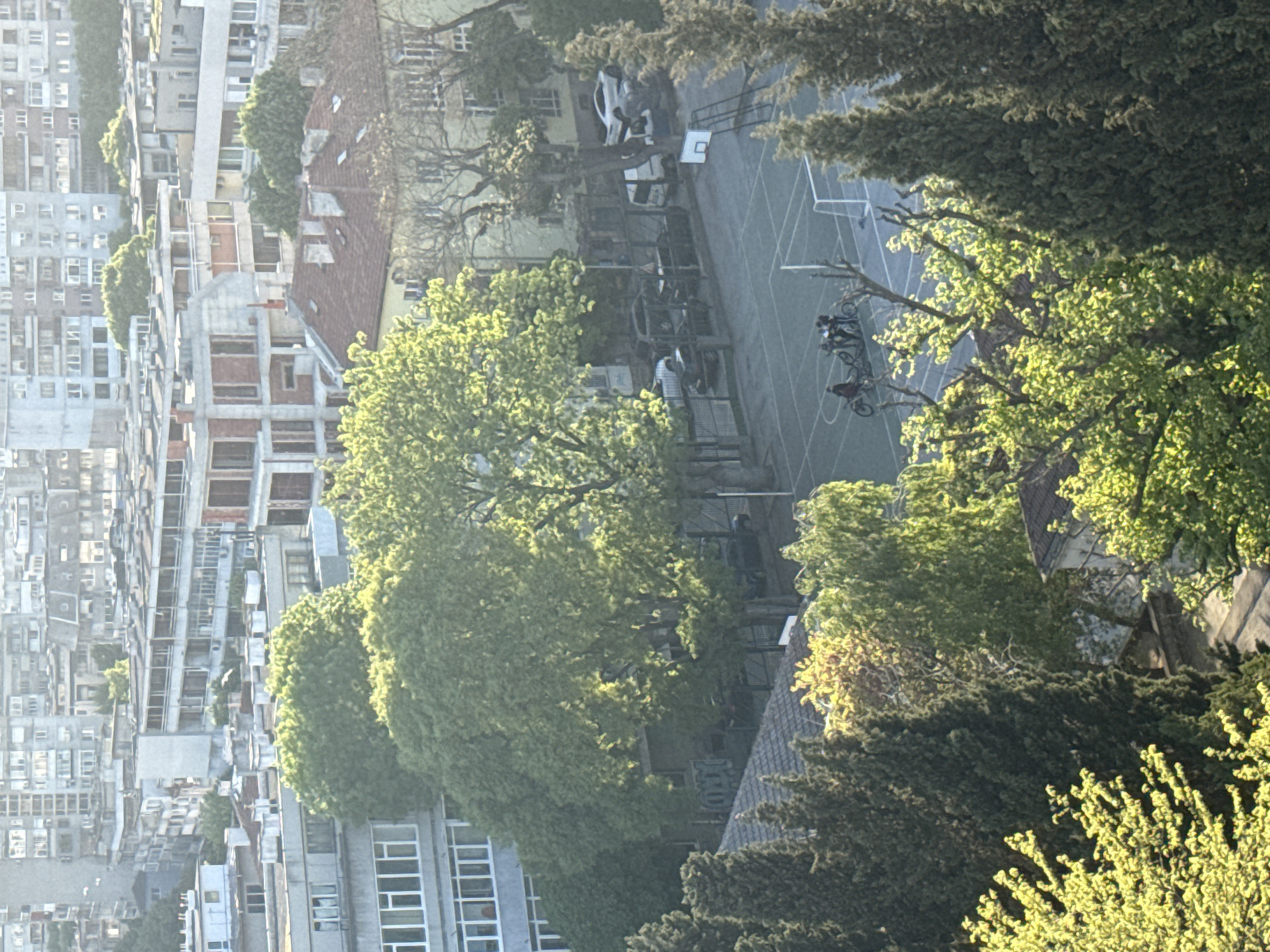 The image shows an elevated view of Varna, Bulgaria. The left side of the image is filled with closely packed, light-colored apartment buildings. To the right, there are trees and greenery. In the middle, partially obscured by trees, there is a gray basketball court with basketball hoops at either end. Bicycles and people are visible on the court. A white car is parked near the basketball court. The environment appears to be urban and possibly a residential area with school facilities.