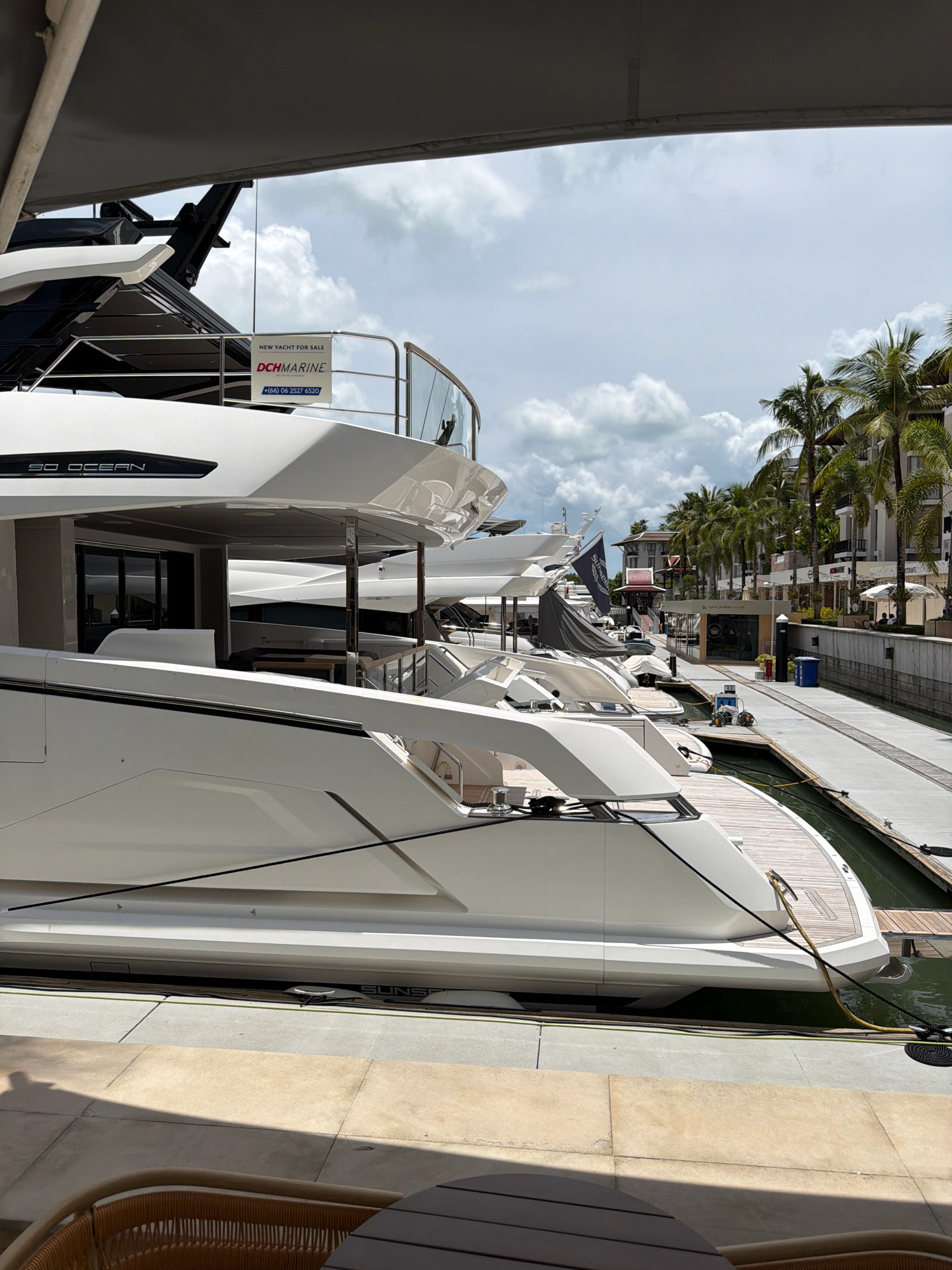 The image depicts a marina in Ko Kaeo, Thailand, under a cloudy sky. Several large, modern white yachts are docked side-by-side. The foreground shows the deck and railing of one of these yachts, with a wooden deck area visible. A sign on a yacht reads "NEW YACHT FOR SALE DCH MARINE +66 06 2527 6520." In the background, palm trees line a walkway that runs alongside the marina, with buildings and shops visible behind them. The overall scene is one of luxury and leisure, with the yachts suggesting a high-end maritime environment.