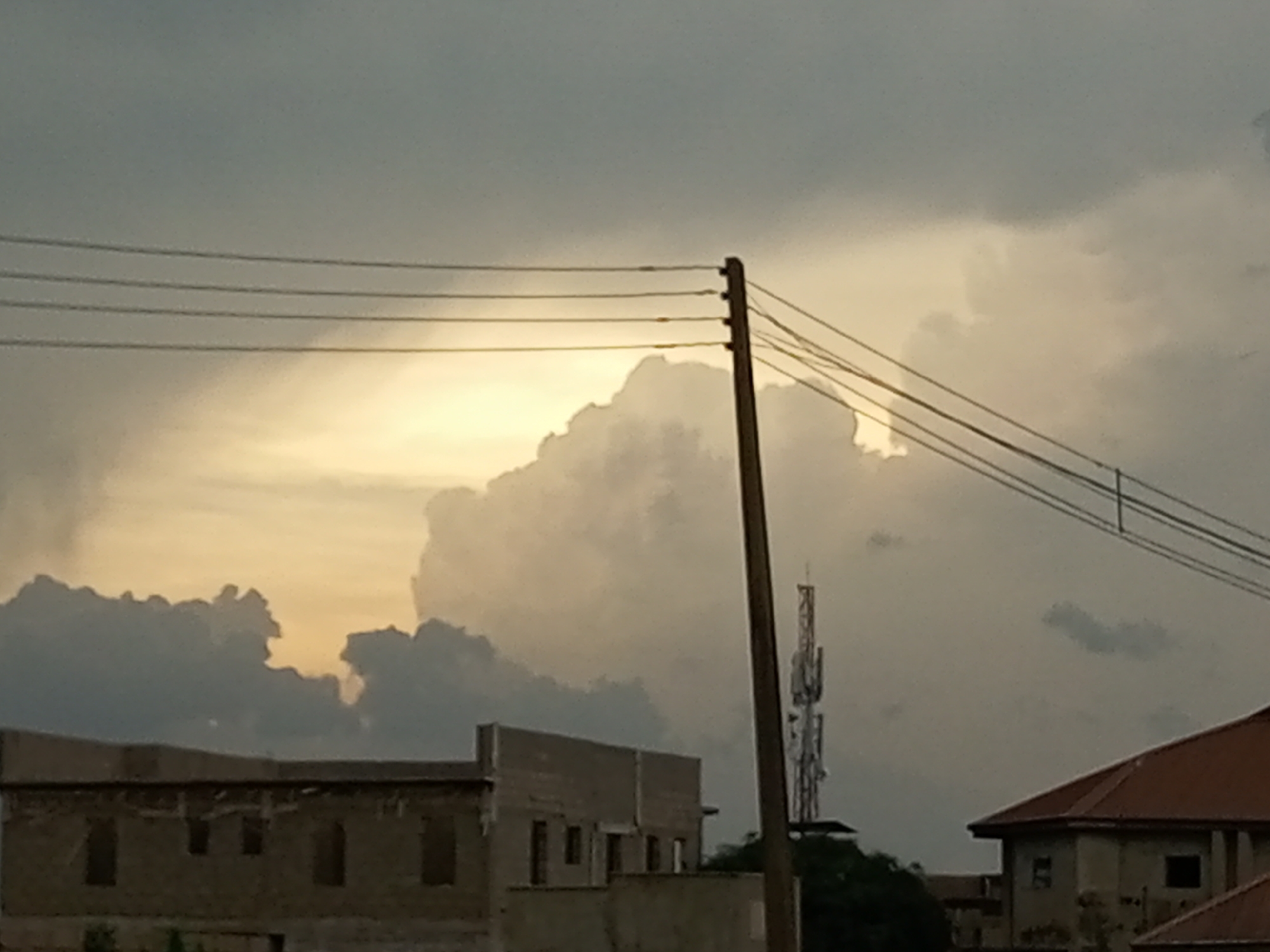 This image depicts a cloudy sky with a dramatic sunlit cloud formation behind a cluster of buildings in the city of Barden Yana, Nigeria. Power lines are strung across the top of the frame, supported by a wooden pole. In the mid-ground, unfinished concrete buildings with dark window openings are visible. To the right of these buildings, a tall telecommunications tower stands against the cloudy sky. Further to the right, the prominent red-tiled roof of another building is visible. The overall impression is one of an urban landscape under a dynamic sky.