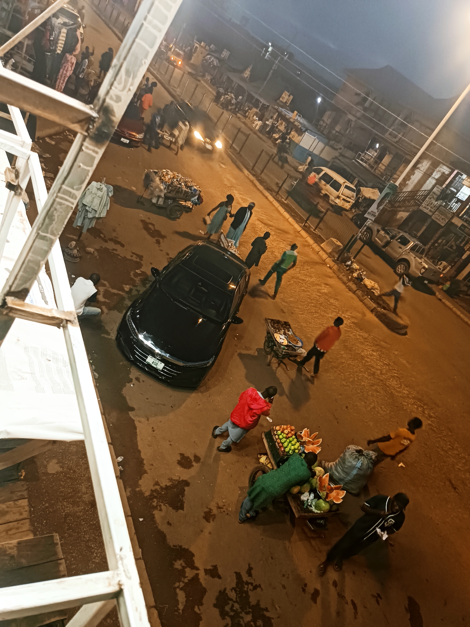 The image captures a busy street scene from a high vantage point, looking down onto the road and its surroundings. The time of day appears to be late evening or night, indicated by the dark sky and prominent artificial lighting, including streetlights and vehicle headlights, which cast warm orange glows on the wet-looking asphalt.
In the foreground, to the left, a white metal scaffolding structure partially obscures the view. Below it, on the street, a black sedan (possibly a Honda Accord, visible grille) is parked or stationary, with a license plate displaying "A5Q". Next to the car, a person in a white t-shirt is sitting on the ground.
The street is bustling with numerous people. Several individuals are seen walking or standing. Notable subjects include a person in a red jacket near the bottom left, and further to the right, a vendor tending to a wooden fruit cart laden with various fruits, including sliced watermelons, apples, and possibly mangoes. Another person in a green garment is also near this cart. Further up the street, another pushcart is heavily loaded with what appears to be discarded items or recyclables.
In the background, more people are visible, along with various vehicles including a white van, a silver pickup truck, and a red car. Buildings line both sides of the street, and a dark metal fence separates the pavement from the road in some sections. The scene suggests a commercial or market area, with people engaged in daily activities such as selling goods, commuting, or socializing. No specific emotions are discernible, as most individuals are captured in motion or engaged in tasks.