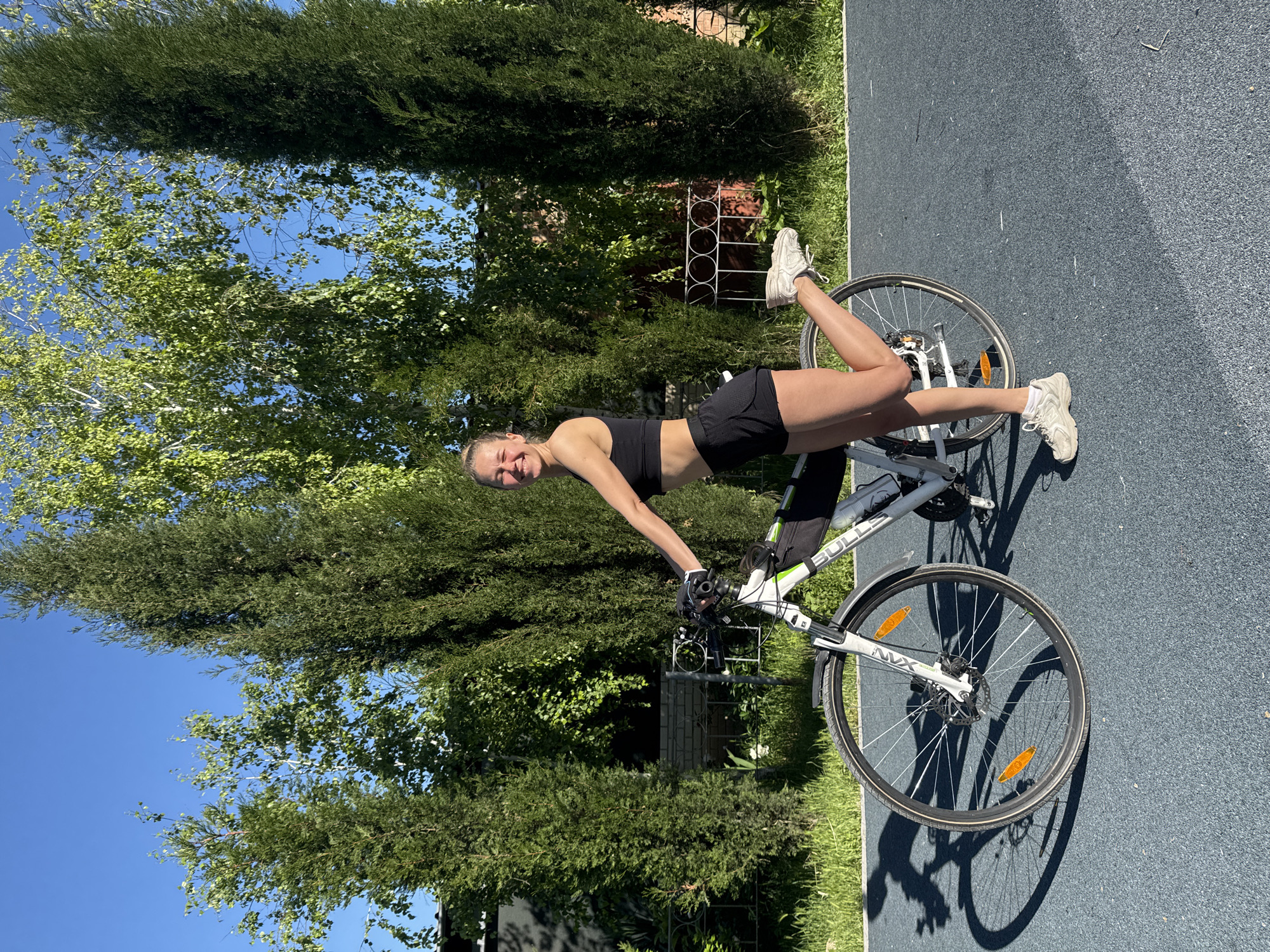 A woman is posing with a white "BULLS" brand bicycle on a paved surface, likely in Мысовское сельское поселение, Ukraine. She is wearing a black sports bra and black shorts, and white sneakers. She is smiling and leaning on the bike with her legs extended onto the bike's front wheel. The background includes greenery, trees, a fence, and a clear blue sky.
