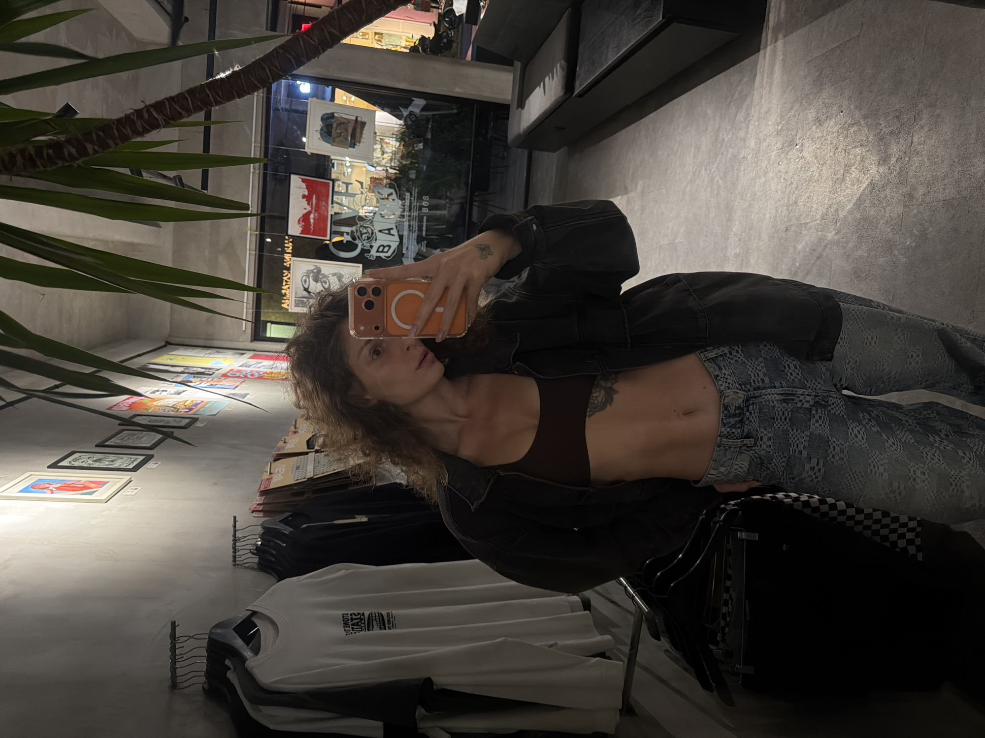 This media file is a first-person, eye-level, vertical photograph taken by a woman in a store. The woman is holding an orange phone in her right hand, taking a selfie. She is wearing a dark jacket over a brown bralette and light-wash patterned jeans. Her curly, brown hair frames her face, and she has a tattoo on her upper arm. Her abdominal muscles are visible.
The background suggests a retail or gallery space. On the left, a large potted plant with long green leaves partially obstructs the view. Behind the woman, the wall displays a collection of framed art pieces, some in white frames and others unframed. The art appears to be posters or prints, with various colors and designs, including red and white. Further in the background, a glass storefront window reveals interior lighting and the reflection of the environment. The floor is a textured concrete.
In the foreground, to the lower right, are stacked t-shirts on display racks. One white t-shirt is clearly visible with black text on it, which reads "STONE" and "20012". Another black garment with a checkered pattern is also visible. The lighting is dim and appears to be artificial, suggesting it is either evening or the interior of a space with limited natural light. The overall mood is casual and contemporary. The location context provided is North Kuta, Indonesia.