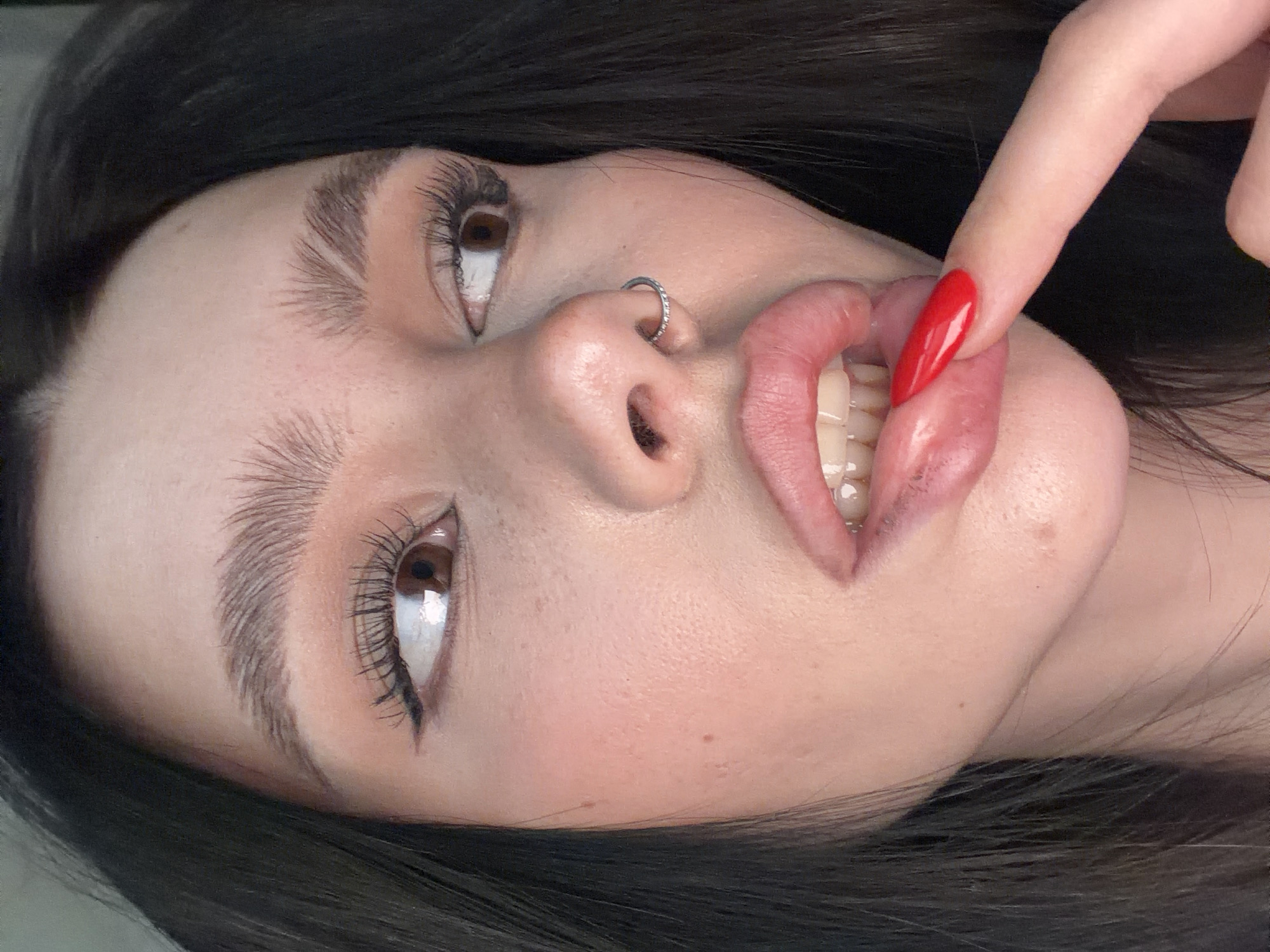 A close-up, low-angle portrait of a female is shown. She has dark hair, fair skin, and full lips. A silver nose ring is visible. She is holding a finger with a red, pointed nail against her lower lip, partially exposing her teeth. The environment is nondescript, offering no indication of Moscow, Russia.