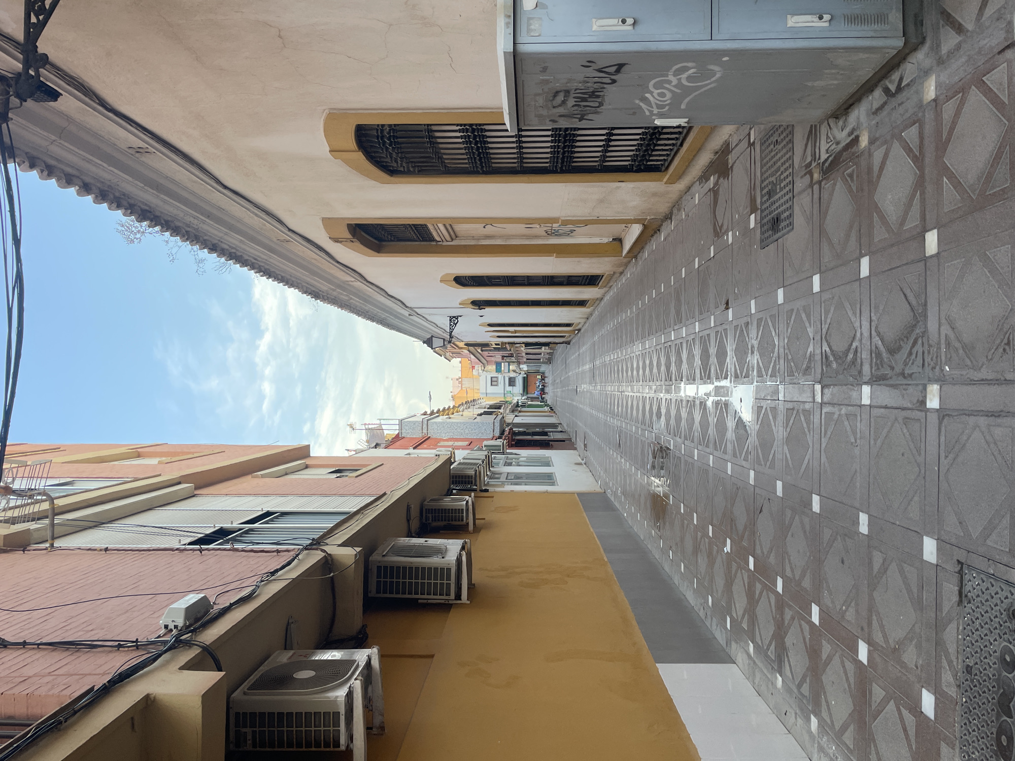 This image shows a narrow street in La Línea de la Concepción, Spain. The street is paved with square tiles featuring a geometric pattern. There are buildings on both sides of the street. The building on the left is painted yellow and has several air conditioning units mounted on its exterior. The building on the right is painted a light beige color and has arched windows with decorative iron bars. A light blue electrical box with graffiti is present on the right side of the street. The street appears to be wet, with puddles reflecting the sky, suggesting recent rain or cleaning. The sky is partly cloudy. In the distance, the street continues, with more buildings and a few people walking.