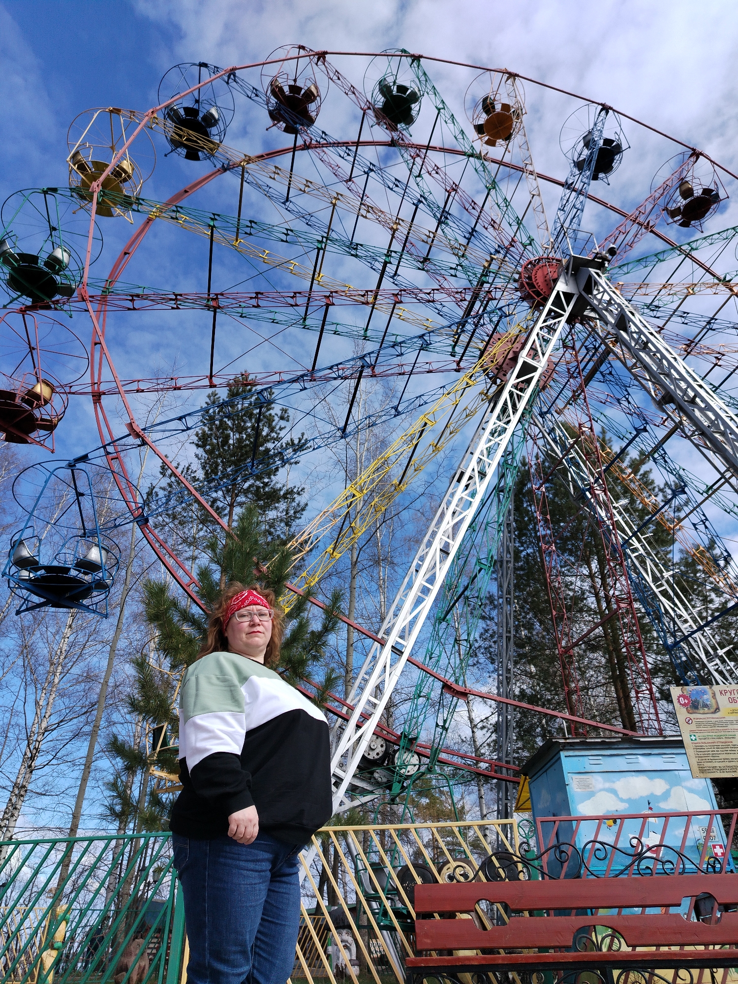 The image depicts a person standing in front of a large Ferris wheel in Chaikovsky, Russia. The Ferris wheel is painted in various colors, with enclosed cabins. The person, wearing a black, white, and green sweatshirt, jeans, and a red bandana, stands to the left of a painted structure and bench. Trees are visible in the background, and the sky is blue with some clouds.